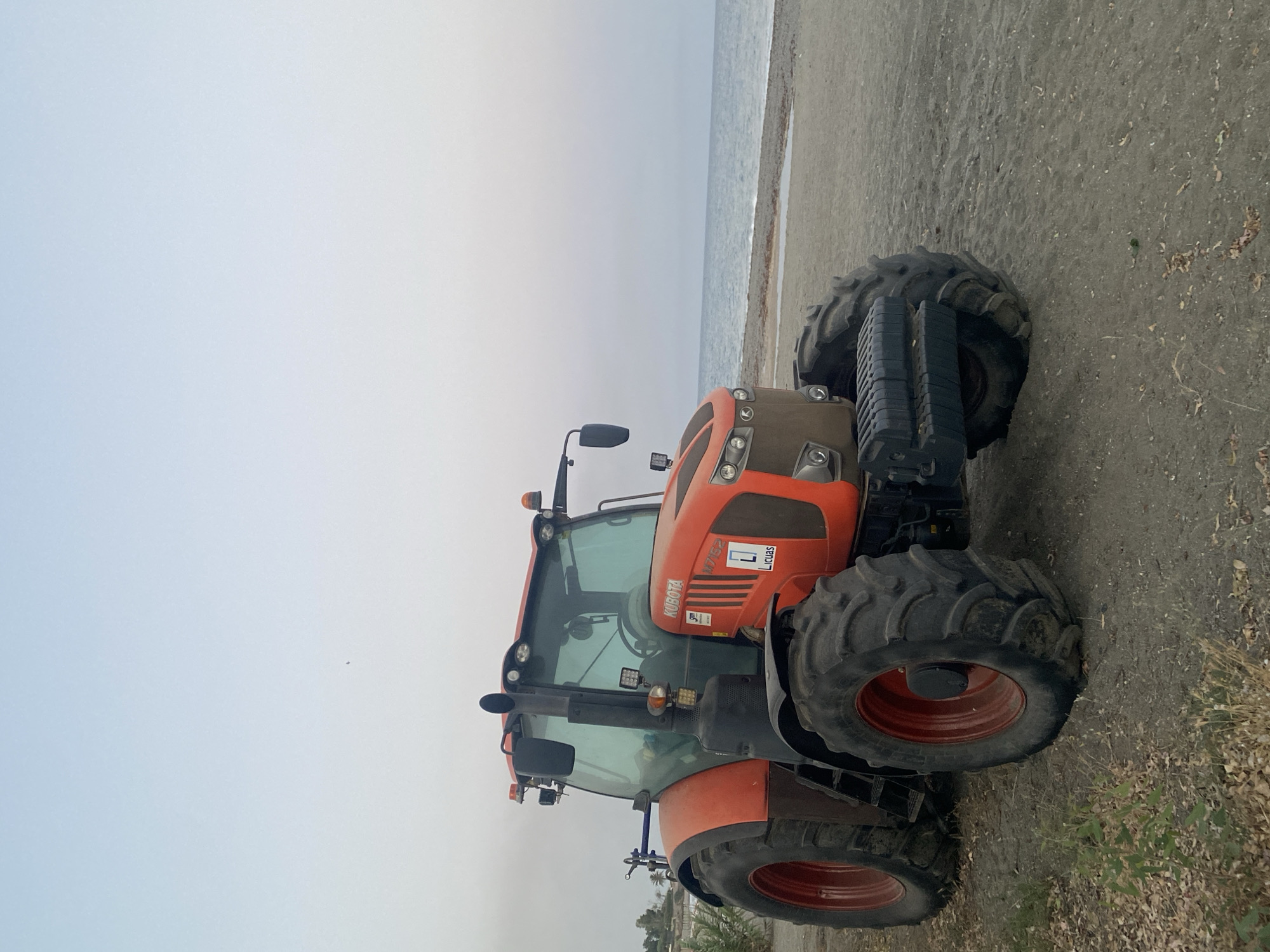 A bright orange Kubota tractor, model M7152, is parked on a sandy beach. The tractor faces the viewer's left, with its front tires deeply embedded in the sand, suggesting it may have been used for beach maintenance or is stuck. The tractor has large, treaded tires with red rims. The cab is visible with tinted windows. There are several lights and warning beacons on the tractor.
In the background, a calm blue sea meets a sandy shoreline. The sky is a pale, hazy blue, indicating it is likely early morning or late evening. The environment appears to be a coastal area, possibly a beach adjacent to a town or resort, given the presence of a tractor. The lighting is soft and diffused. While the image is taken in Estepona, Spain, there are no specific identifiable landmarks within the frame to pinpoint the exact location within the city. The overall scene is quiet and still.