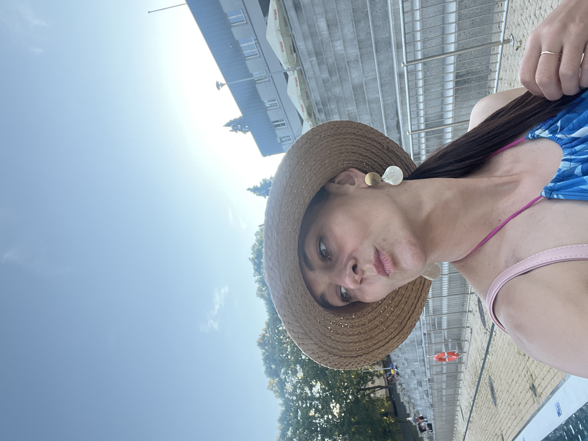 A woman with dark hair and a straw hat is posing for a selfie outdoors. She is wearing a blue and white patterned swimsuit and a pink strap. She has on large, unique earrings. Behind her, there are tiered seating areas with railings, a building with windows, and trees. The sky is clear and blue with some very faint clouds. A life preserver hangs on a railing near the seating area. The scene appears to be at a public pool or outdoor recreational facility.