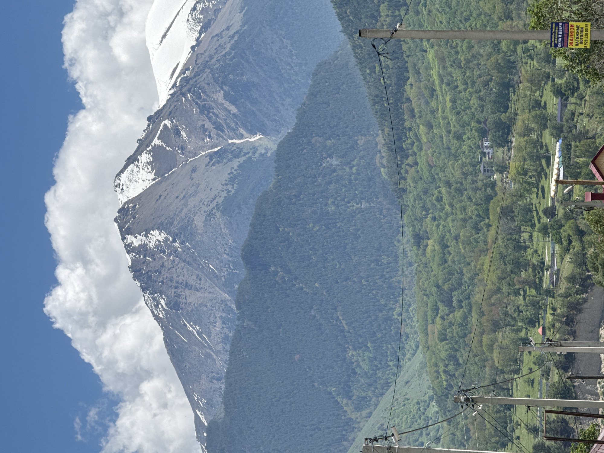 The image depicts a vast mountain landscape dominated by a snow-capped peak under a bright blue sky with fluffy white clouds. The foreground is occupied by a densely forested mountainside, with lush green trees covering the slopes. Several utility poles with electrical wires are visible in the lower portion of the image, suggesting human presence and infrastructure. In the middle ground, a small settlement with houses and buildings is nestled amongst the trees. The overall scene conveys a sense of natural beauty and tranquility, with a juxtaposition of rugged mountains and verdant valleys. The signage on the pole in the lower right corner of the image indicates that the location is Teberda, Russia.