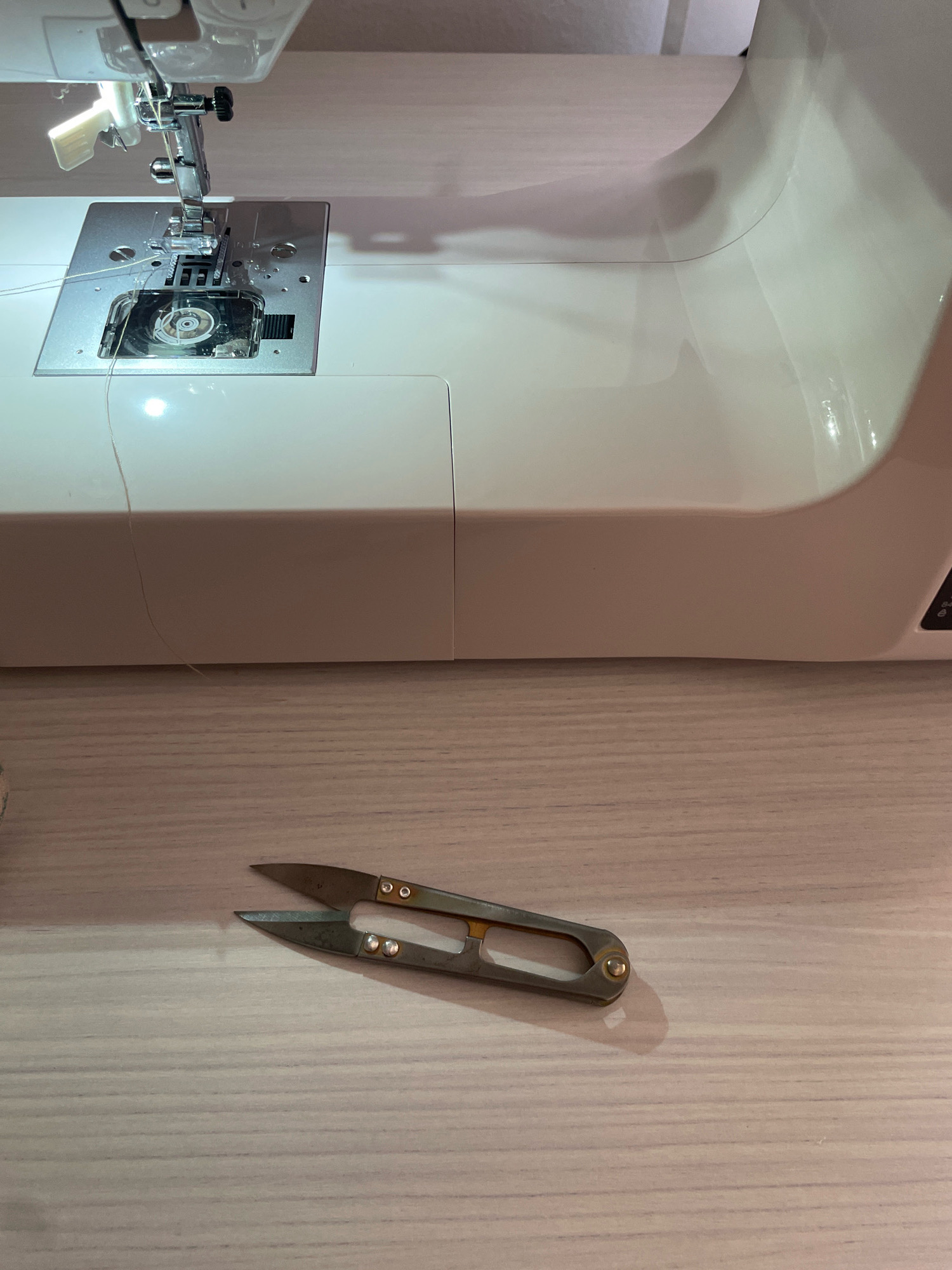 A close-up, eye-level shot shows a sewing machine and a pair of thread snips on a light-colored wooden surface. The sewing machine is a white, modern model, with its needle and presser foot visible and engaged with the fabric plate. A thin, light-colored thread is visible running from the needle and disappearing into the machine. The thread snips are in the foreground, open and resting on the wooden surface. They are made of metal with gold accents and appear to be used. The lighting in the image is warm and bright, creating soft shadows. This image was taken in Atarfe, Spain.