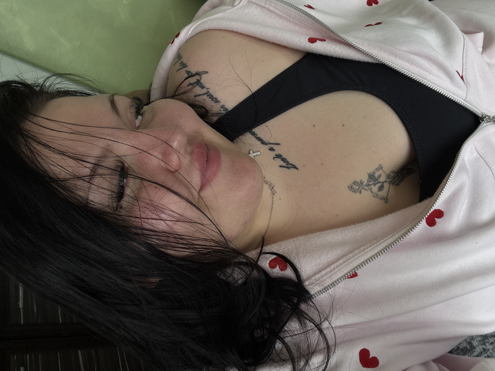 A woman lies at an angle, wearing a black top and a pink zip-up hoodie with red hearts. She has dark hair and tattoos on her chest and shoulder. A silver necklace is visible. The background includes green and dark brown surfaces. Location information not discernible.