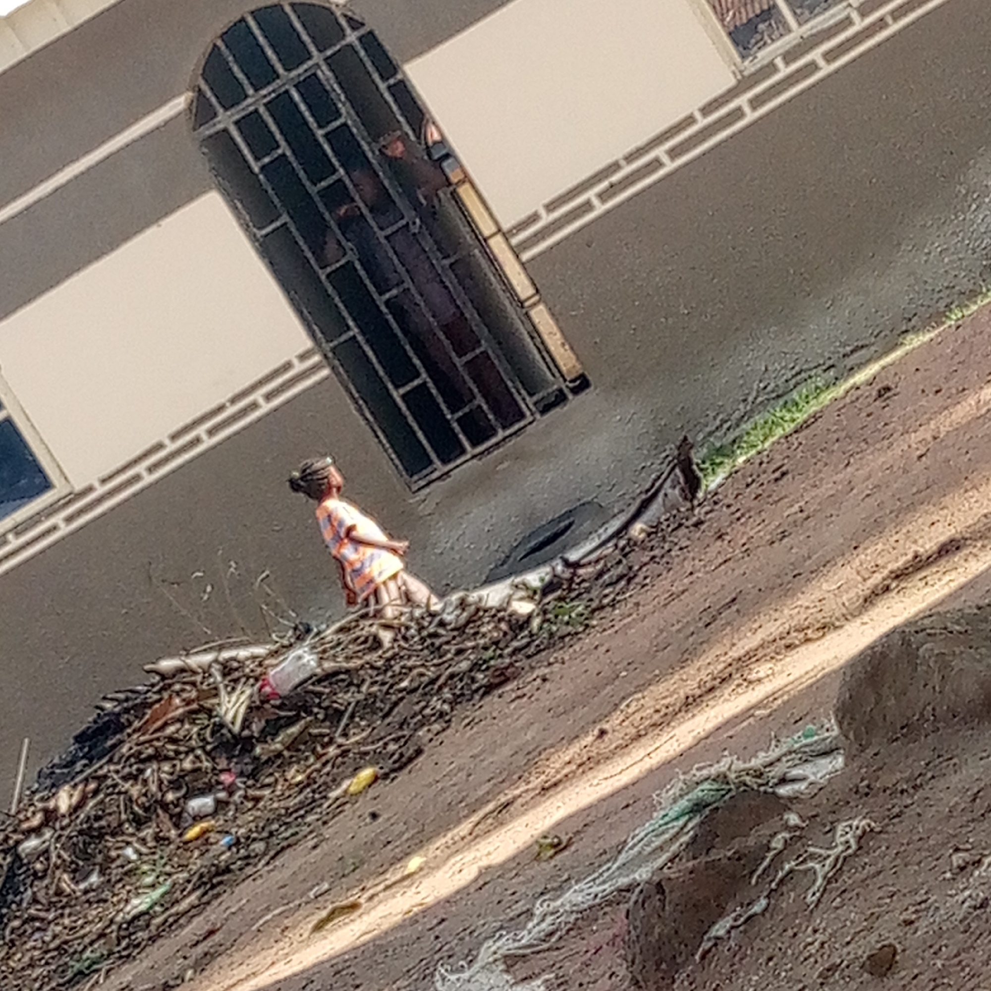 A young African girl wearing a peach and orange patterned shirt and brown pants stands on a pile of trash next to a gray building in Mararaba, Nigeria. The building has a unique arched window with black bars. The trash heap consists of plastic bottles, sticks, and other debris. The ground is a mix of dirt and trash.