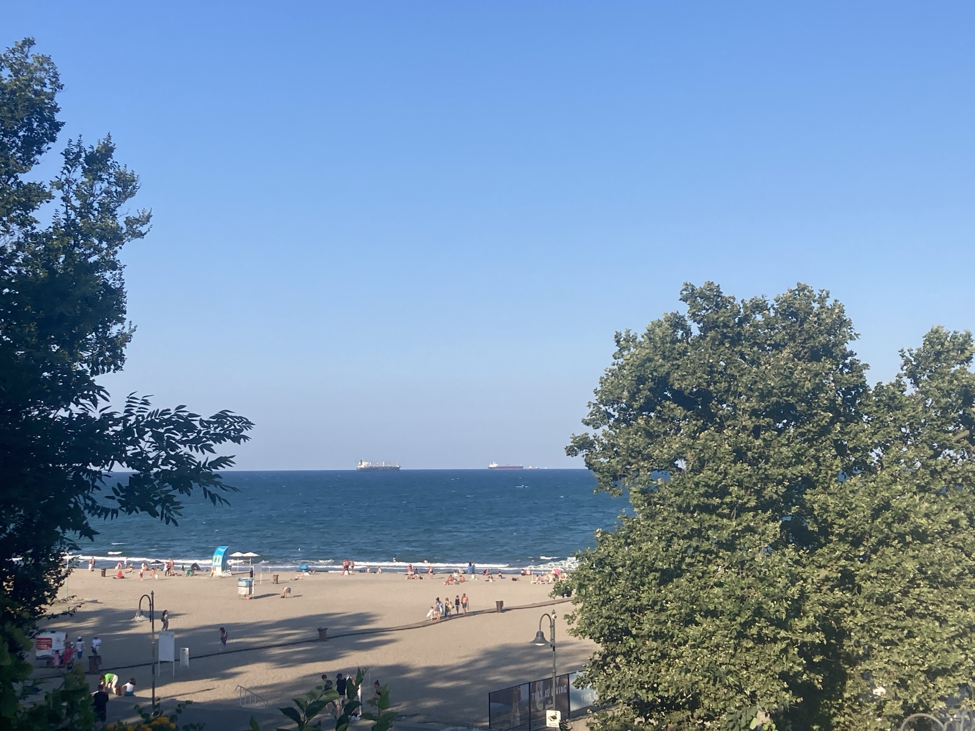This is an outdoor, eye-level, wide shot of a crowded beach scene in Burgas, Bulgaria. The image captures the sea, beach, and a portion of a promenade or walkway. The sky is clear and blue. In the foreground, two large trees frame the view. The left tree is dark green and bushy, its leaves creating a partial silhouette. The right tree is also green and leafy, its branches extending towards the center of the frame.
The beach itself is light brown sand and is populated by numerous people. Some are sunbathing, others are walking, and some are sitting. There are several beach umbrellas and chairs scattered across the sand. A blue and white beach tent is visible.
The sea is a deep blue with moderate waves. Several large cargo ships are visible on the horizon, indicating maritime activity.
The promenade area shows some people walking. There are lampposts, trash bins, and some signage. The overall atmosphere suggests a warm, sunny day at a popular seaside location. The presence of people, beach amenities, and ships points to a typical day at a coastal resort area.