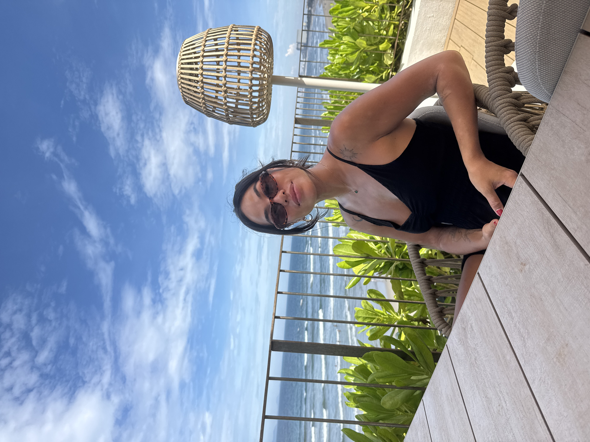 A woman wearing sunglasses and a black tank top sits in a wicker chair at an outdoor table. Behind her is a metal railing, green vegetation, and a view of the ocean and beach. A wicker lampshade hangs above. The sky is blue with scattered clouds. The location is likely a patio or balcony overlooking the beach. The city is Đà Nẵng, Vietnam.