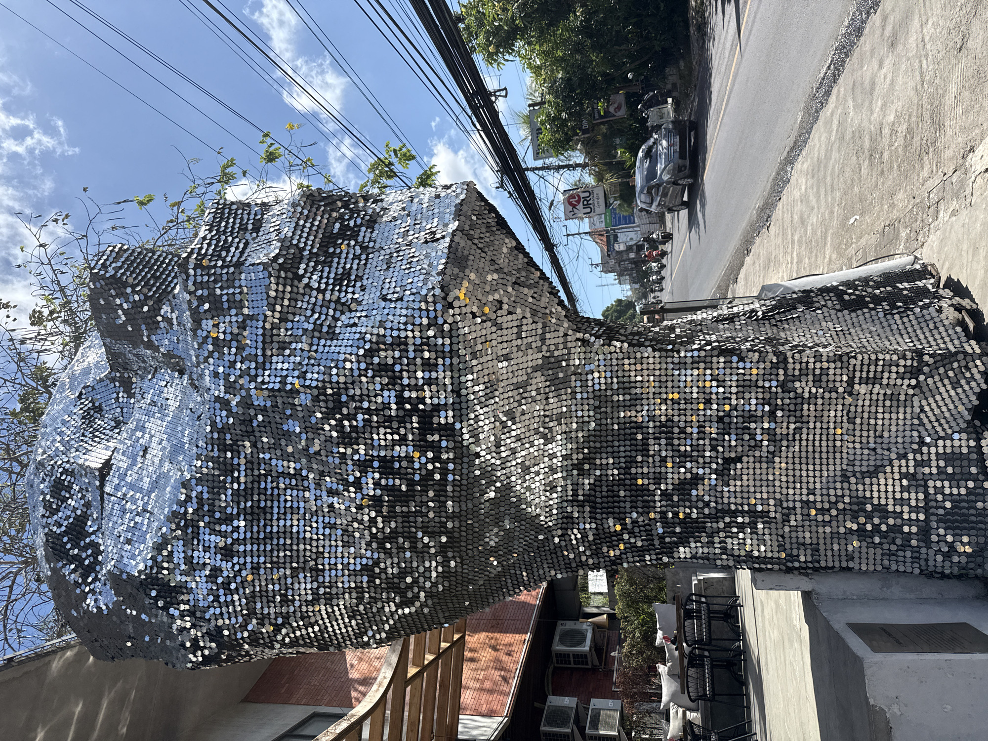 In Kuta Selatan, Indonesia, a sequined sculpture, possibly depicting a stylized head, stands on a concrete sidewalk next to a street. The sculpture is covered in numerous small, reflective silver sequins that catch the sunlight. Trees with sparse leaves and yellow flowers grow from the top of the sculpture. To the left of the sculpture, a building with a brick facade and awnings is visible, with outdoor seating, tables, and chairs, as well as several air conditioning units mounted on the wall. A street with passing cars and motorcycles stretches into the distance, lined with trees and utility wires overhead. A sign for "URU RAMEN & SUSHI" is visible on the right side of the street. The sky is blue with scattered white clouds.