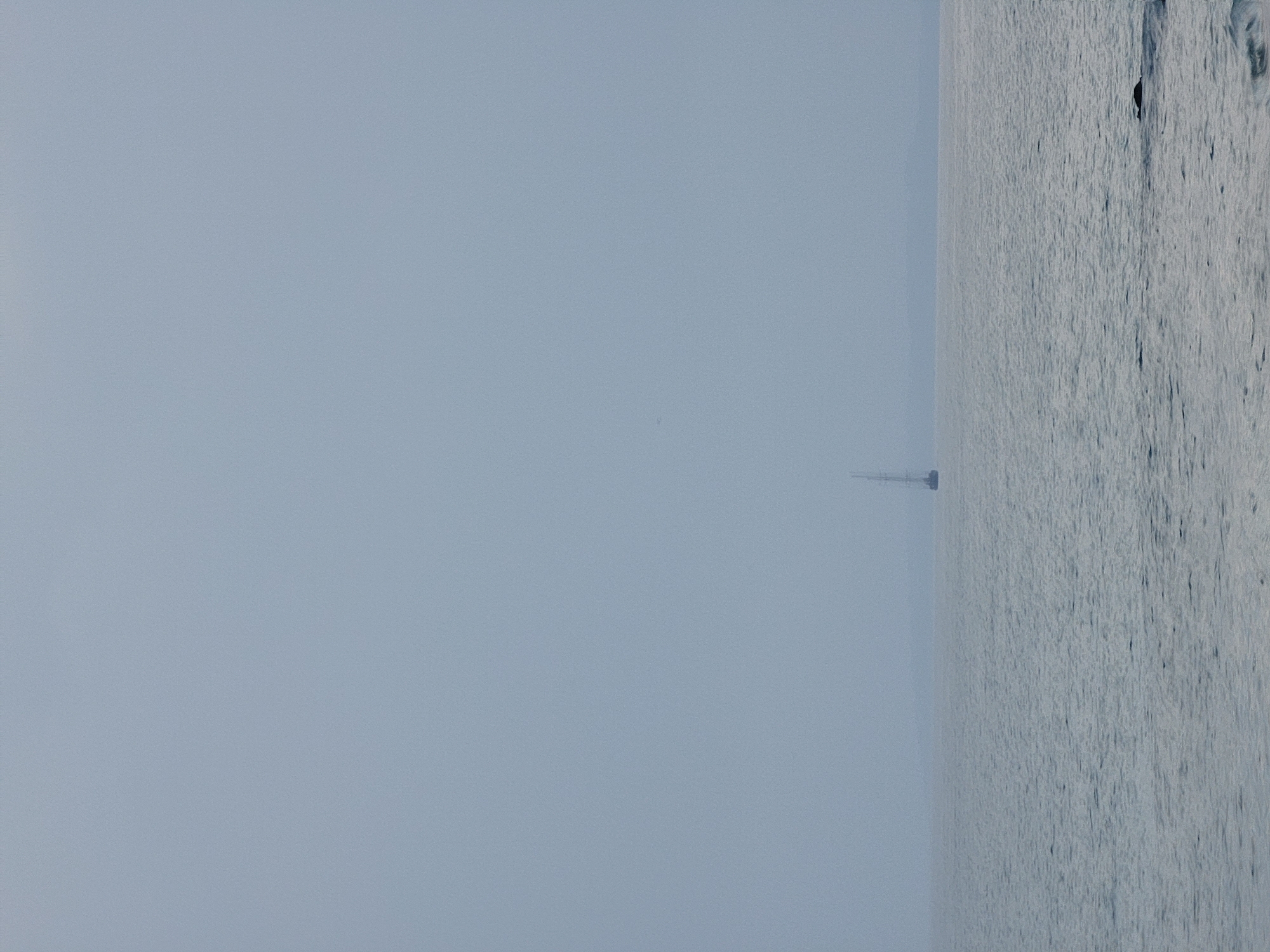 A distant, tall-masted sailboat is visible on the horizon of a calm sea under a hazy, overcast sky. The water is a muted blue-grey, with subtle ripples. The sky is a uniform pale grey, obscuring any details of the distant landmass. A dark shape, possibly a rock or a small island, is partially submerged in the water in the foreground to the right. The scene evokes a sense of quiet solitude. The image was taken off the coast of Quarteira, Portugal.