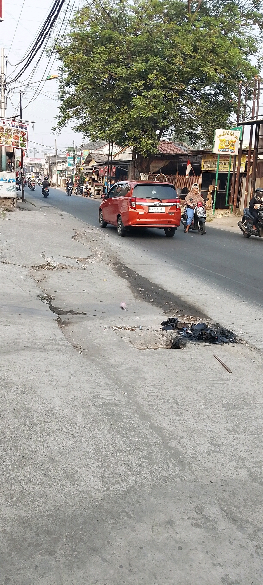 The image depicts a street scene in Satriamekar, Indonesia, on a sunny day. The street is paved with concrete and shows signs of wear, including cracks and a visible pothole filled with debris. To the left of the street, a sidewalk is present, bordered by a line of shops and buildings. A large tree with dense green foliage stands prominently in the middle ground, casting shadows on the surroundings. Power lines crisscross the sky above.
Several vehicles are visible on the road. An orange car is in the foreground, heading away from the viewer. A woman on a scooter is also moving away, with a passenger behind her. Several other motorcycles are present further down the street, some parked and others in motion, indicating a typical urban traffic flow.
Commercial signage is present on the buildings, with one sign on the left displaying food items and the text "NASI UDUK." Another sign on the right, featuring cartoon characters, reads "KEDAI SEDJAK & SOP IOO."
The environment appears to be a bustling commercial area with a mix of residential and business establishments. The presence of litter, including a pink plastic cup and black garbage bags in the pothole, suggests an ongoing urban environment. The overall atmosphere is that of a busy street in a Southeast Asian city.