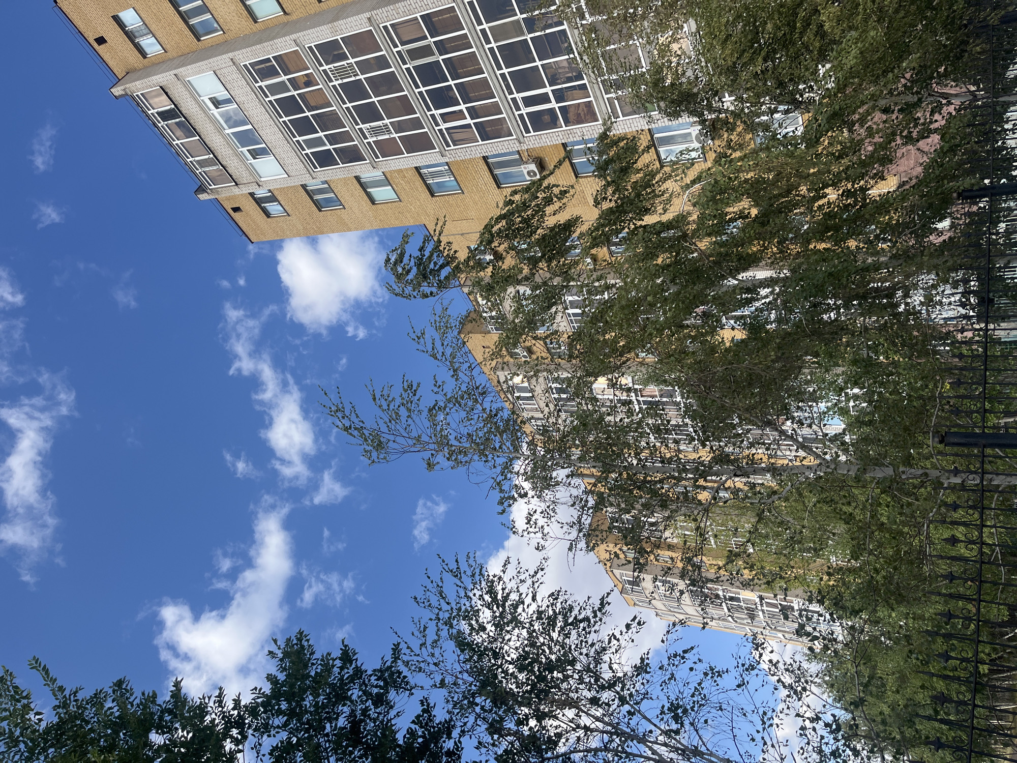 The image depicts a view of residential buildings against a bright blue sky with scattered white clouds. The buildings appear to be modern, with yellow and grey brick facades, multiple stories, and numerous windows, many of which are enclosed balconies with glass fronts. Trees with green foliage are in the foreground, partially obscuring the buildings. A black wrought-iron fence runs along the bottom of the image. The scene is likely from a city like Astana, Kazakhstan, given the architectural style and the presence of such apartment blocks. There are no people or animals visible in the image. The overall impression is one of an urban residential area on a clear day.