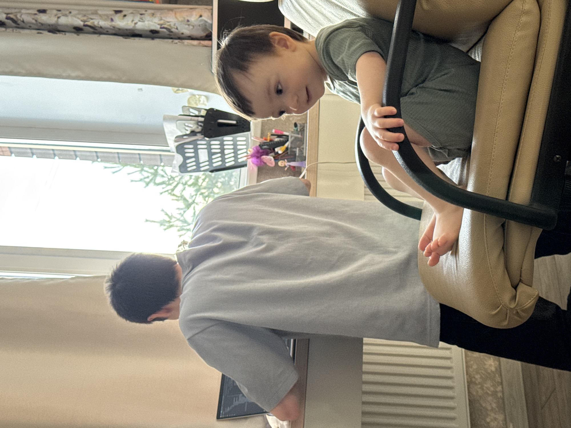 A young boy wearing a green outfit is sitting in a beige office chair, looking down at an older boy or man who is standing at a desk. The desk is next to a window and contains a computer, stationery items and a basket. The location is unspecified.