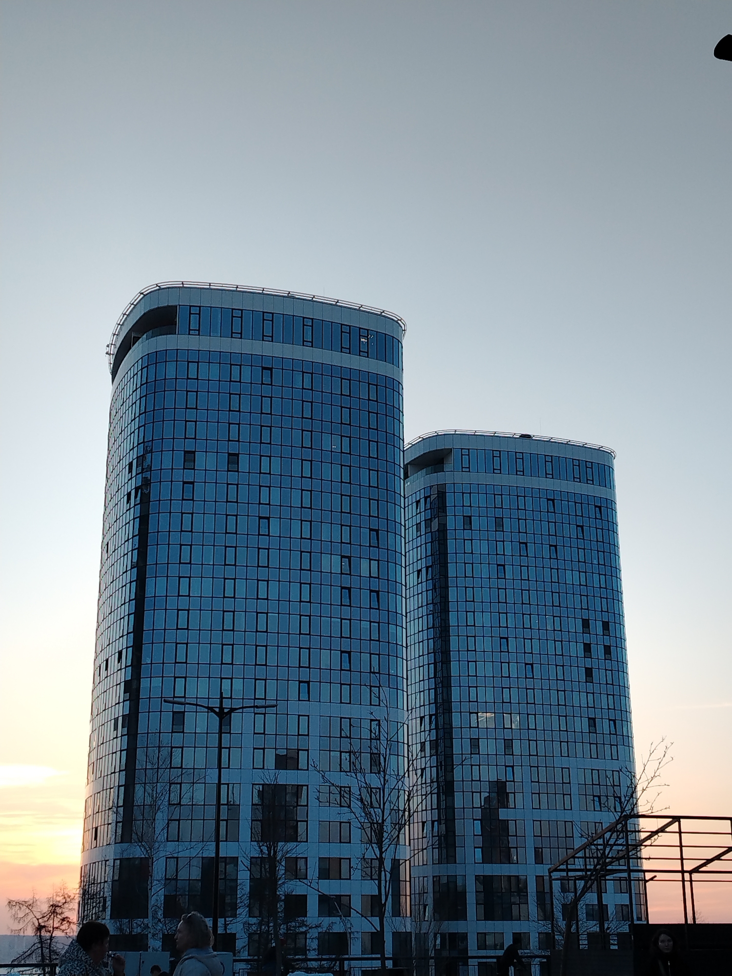 The image displays two prominent, tall, cylindrical-shaped high-rise buildings with blue-tinted glass facades, situated in the city of Izhevsk, Russia. The sky overhead transitions from a light blue-grey at the zenith to a subtle pinkish-orange near the horizon on the left, indicating either dawn or dusk. Each building exhibits a grid pattern of numerous square window panes, with several individual windows appearing partially ajar on both structures. In the foreground, below the buildings, bare deciduous trees are visible, along with a dark, multi-headed street lamp post silhouetted against the structures. Several partially obscured human figures are present in the lower left portion of the foreground; one individual with dark hair is seen looking towards the right, and another with lighter hair is partially visible looking towards the left. A dark, angular metal structure, possibly a railing or framework, is visible in the lower right foreground.