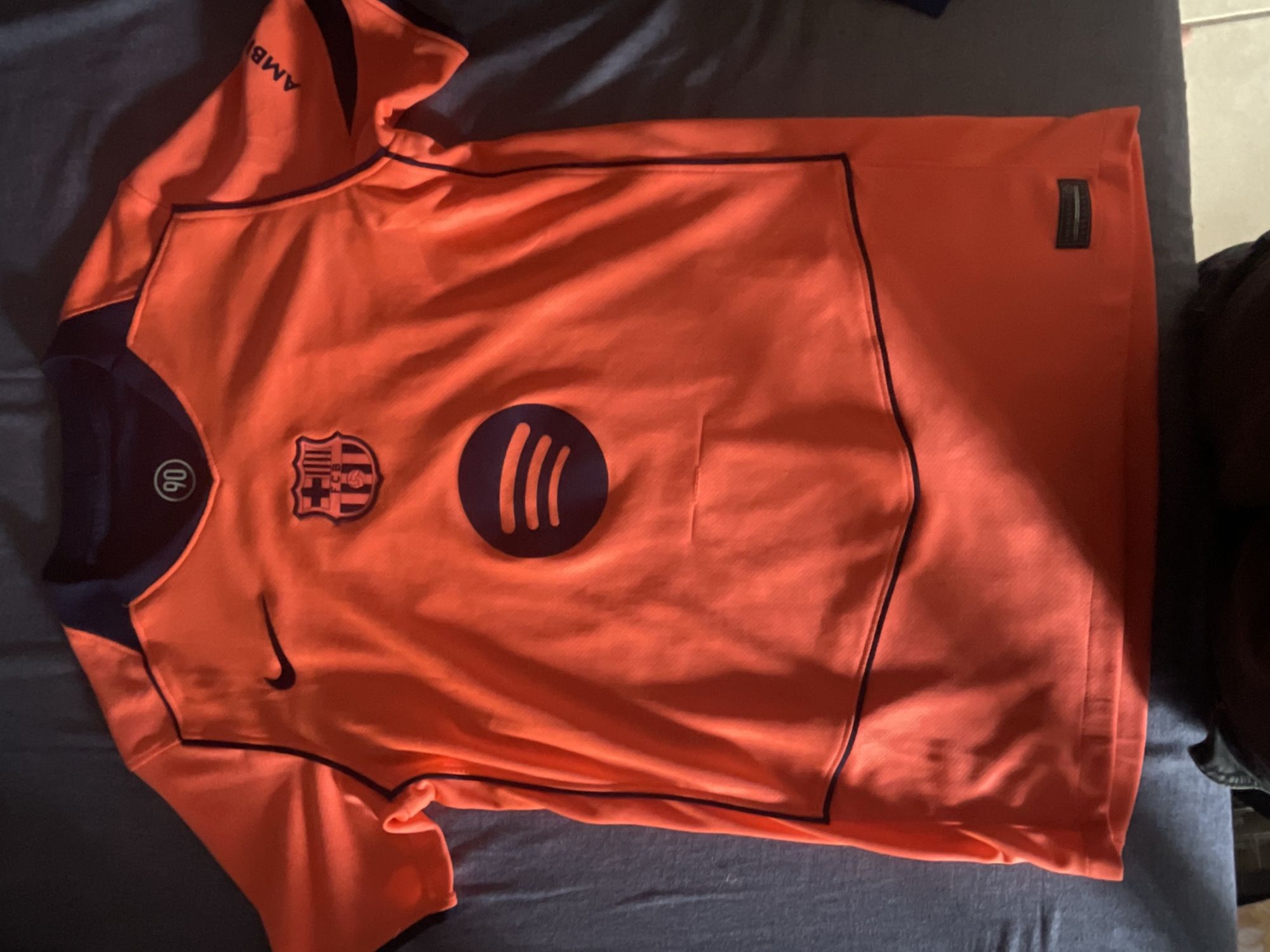 A folded, bright orange, short-sleeved soccer jersey is displayed flat on a dark gray, textured surface. The jersey features navy blue accents around the collar, shoulders, and down the sides. Prominently displayed on the chest is the FC Barcelona crest in navy blue and red, with a Nike logo in black to the left. Below the crest, a large navy blue Spotify logo is centered on the jersey. Text, "AMB," is visible on the right sleeve in navy blue. A small circular tag with "90" is present near the neckline on the left side. A rectangular tag with faint white text and a silver outline is attached to the lower right hem. The jersey appears to be made of a performance fabric with a subtle textured pattern.
