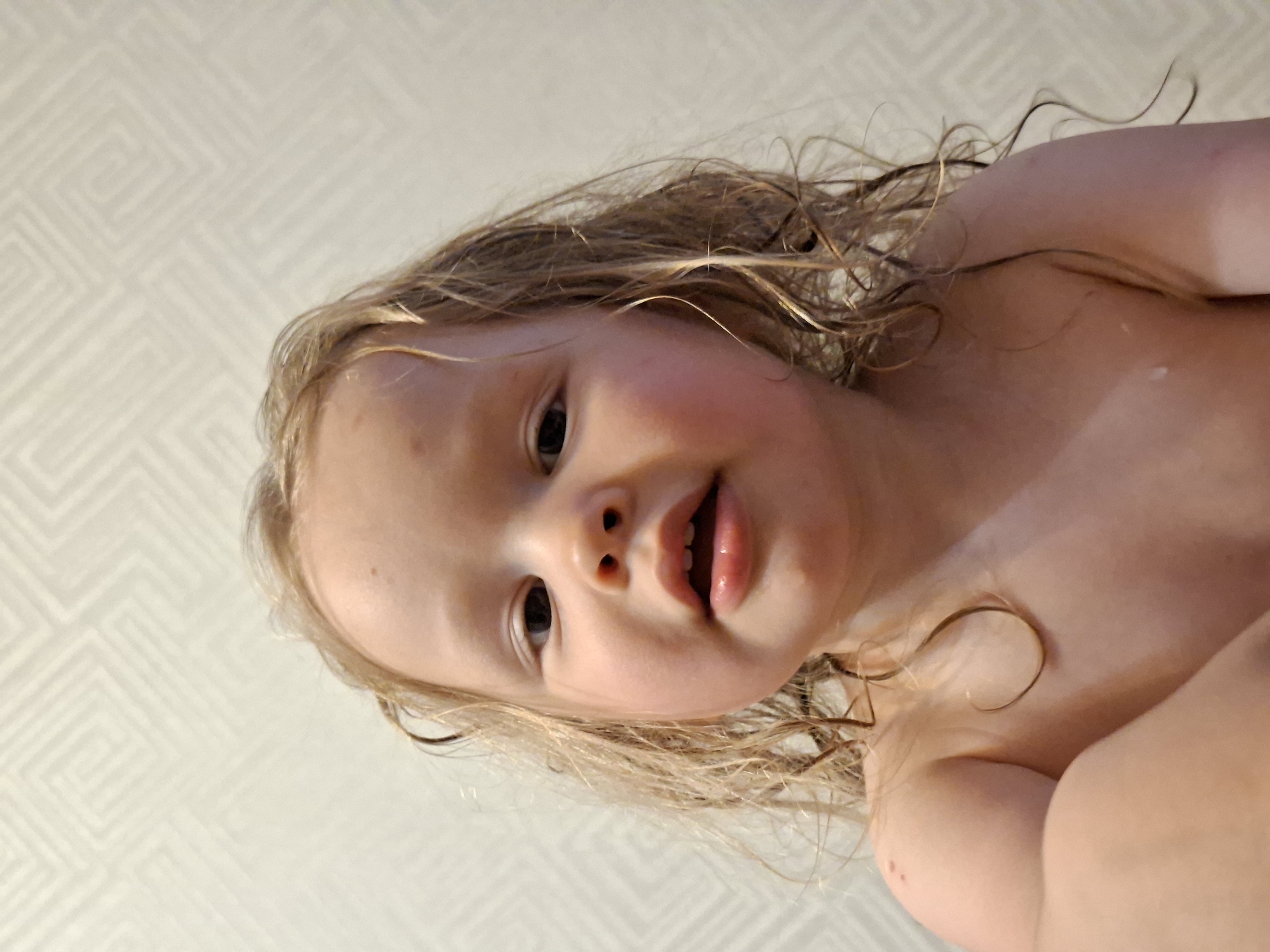 A close-up shot focuses on a young child with fair skin and damp, light-colored hair. The child looks up at the camera with an open mouth. A geometric pattern is visible in the background. The city and state are unknown.