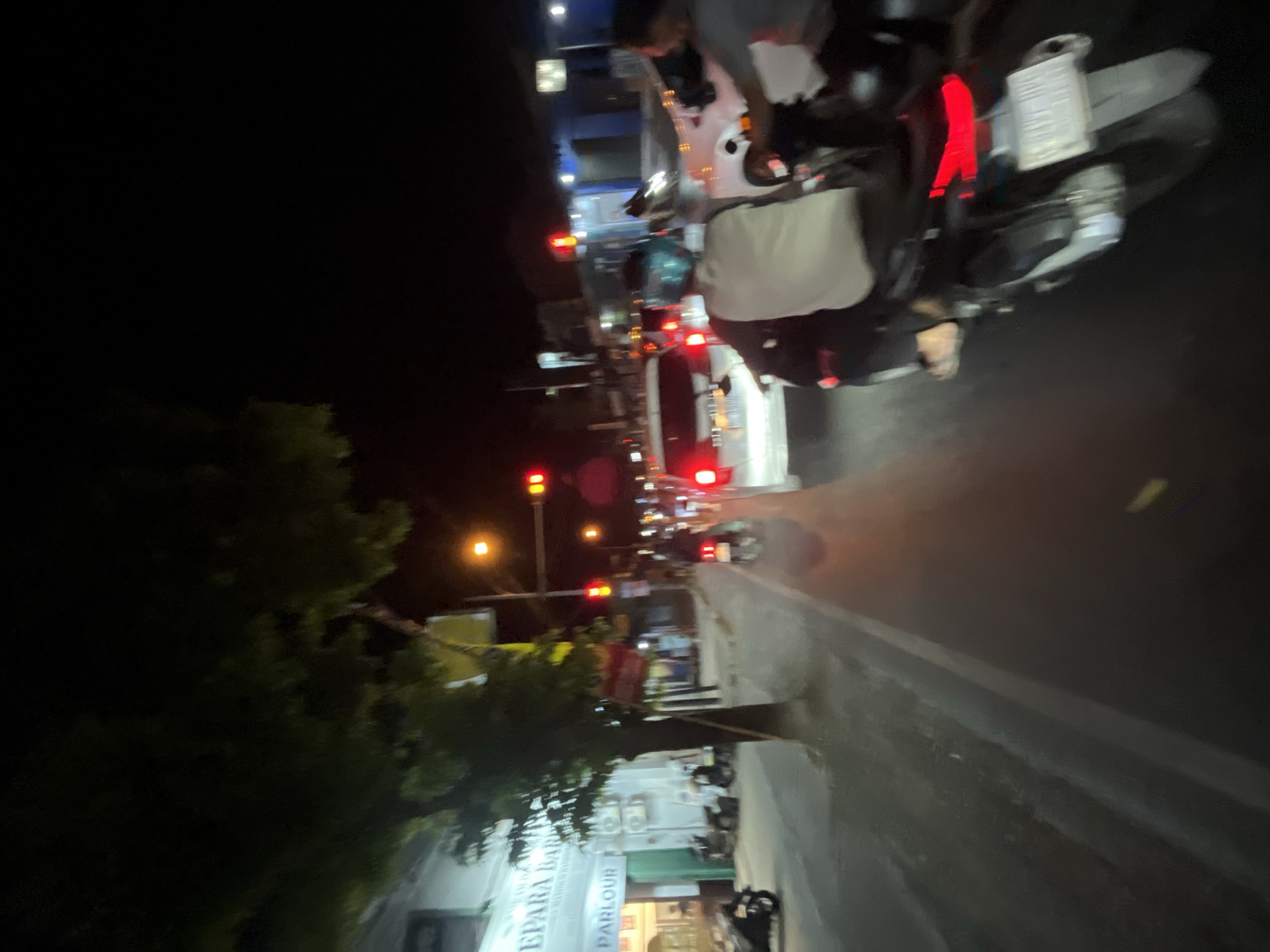 This image depicts a street scene at night in Kalinyamatan, Indonesia, likely a traffic intersection with functioning traffic lights. A white car is stopped at the red light, as are several motorcycles. On the right side of the road, a person rides a motorcycle, wearing a light-colored shirt and a blue helmet. Another person is visible on the back of this motorcycle. In the foreground, another motorcycle is partially visible. To the left, a building with illuminated signage can be seen, with a tree in front of it. The street is wet, suggesting recent rain or a humid environment. The overall atmosphere is one of a typical busy urban street at night.