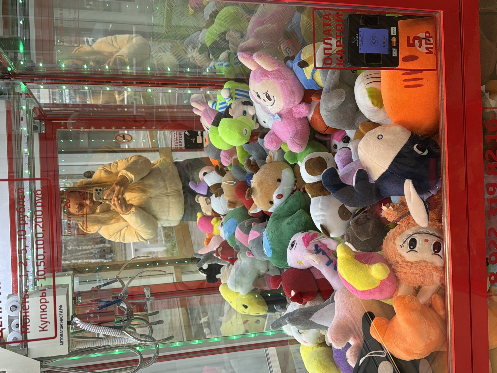 This is an eye-level, slightly downward-angled shot inside a claw machine filled with numerous plush toys, located in Votkinsk, Russia. The machine's glass panels reflect a person, likely the photographer, wearing a light-colored jacket and holding a smartphone, capturing the scene. Green LED lights line the interior edges of the machine, creating a vibrant effect.
The main subjects are the diverse collection of plush toys, packed densely together. They include various animals like rabbits, foxes, a shark, and characters in different colors and sizes, such as a pink bunny, an orange shaggy character, and a black-and-white dolphin-like toy.
The claw mechanism is visible in the lower left, hanging ready for play. The machine features several payment options. Text on the left panel states "Монеты, 10 рублей" (Coins, 10 rubles) and "Купюры - 10, 50, 100, 200 руб." (Banknotes - 10, 50, 100, 200 rubles). Below it, a website address "АВТОМАТЗАПЧАСТИ.РФ" (AVTOMATZAPCHASTI.RF) is printed. A sticker further specifies "ЦЕНА ИГРЫ 20" (Game price 20), and mentions accepted coins "2, 5, 10" and banknotes "10, 50, 100 руб".
On the right side of the machine, a digital card payment terminal is installed, featuring the "vendista" brand and displaying "ПОДВЕСИТЕ КАРТУ" (Tap Card) and "100 РУБ" (100 rubles), with logos for VISA, MasterCard, and МИР (MIR). Above this terminal, "ОПЛАТА КАРТОЙ" (Card Payment) is written, along with "5 ИГР" (5 games), potentially indicating a multi-play option. A phone number starting with "8 982 16" is partially visible on the red frame. The activity taking place is the observation or preparation to play this arcade game. The setting is likely an indoor public space.