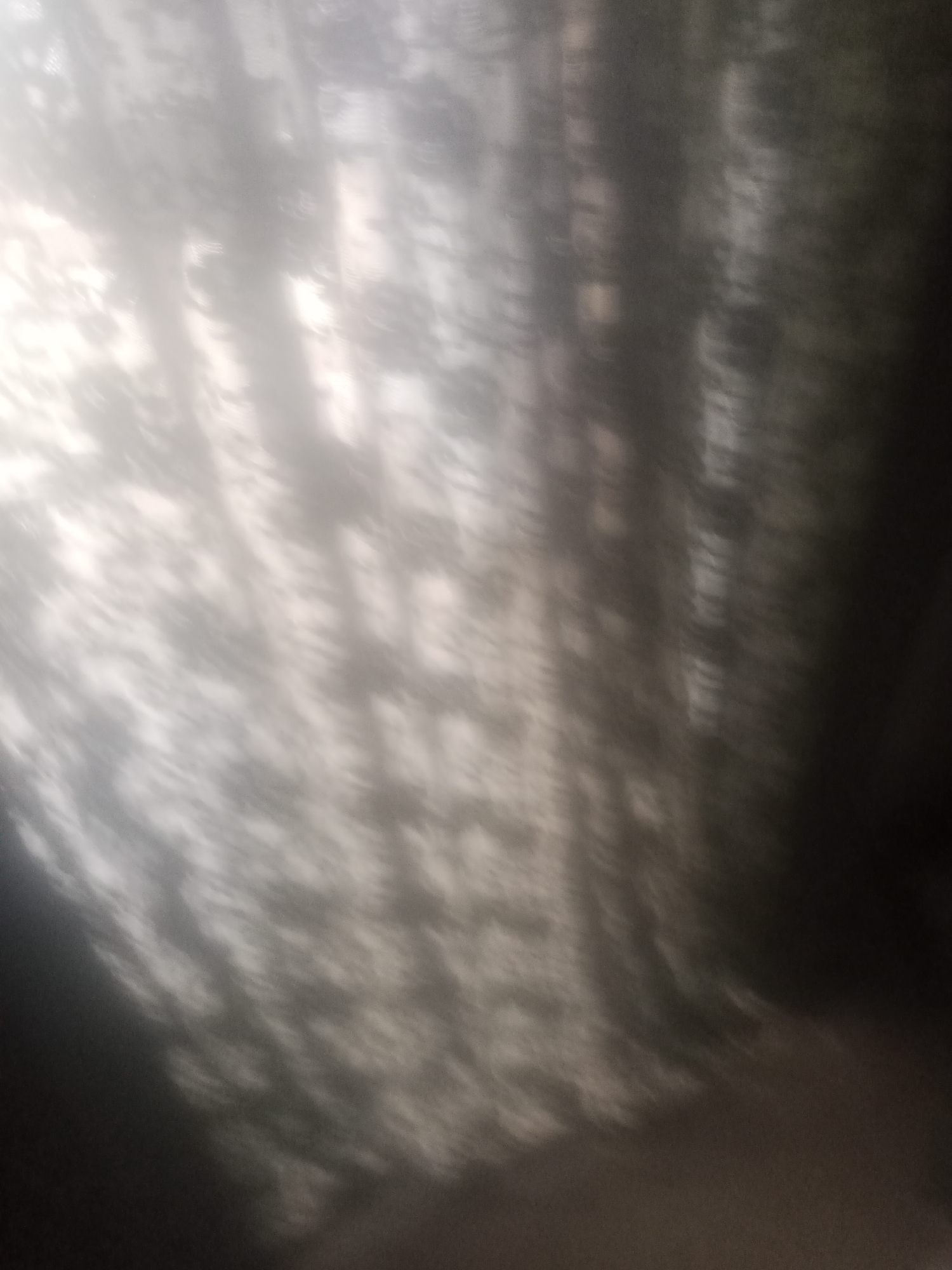 The image is a close-up, out-of-focus view of a patterned curtain. The pattern appears to be a floral or lace design, with light and dark areas creating a textured effect. The lighting suggests the sun is shining through the curtain, casting shadows and highlights that contribute to the abstract appearance. The colors are muted, ranging from light gray to dark gray and white. There are no people, animals, or discernible objects in the scene other than the curtain itself. The image does not provide any information about a specific location or city.