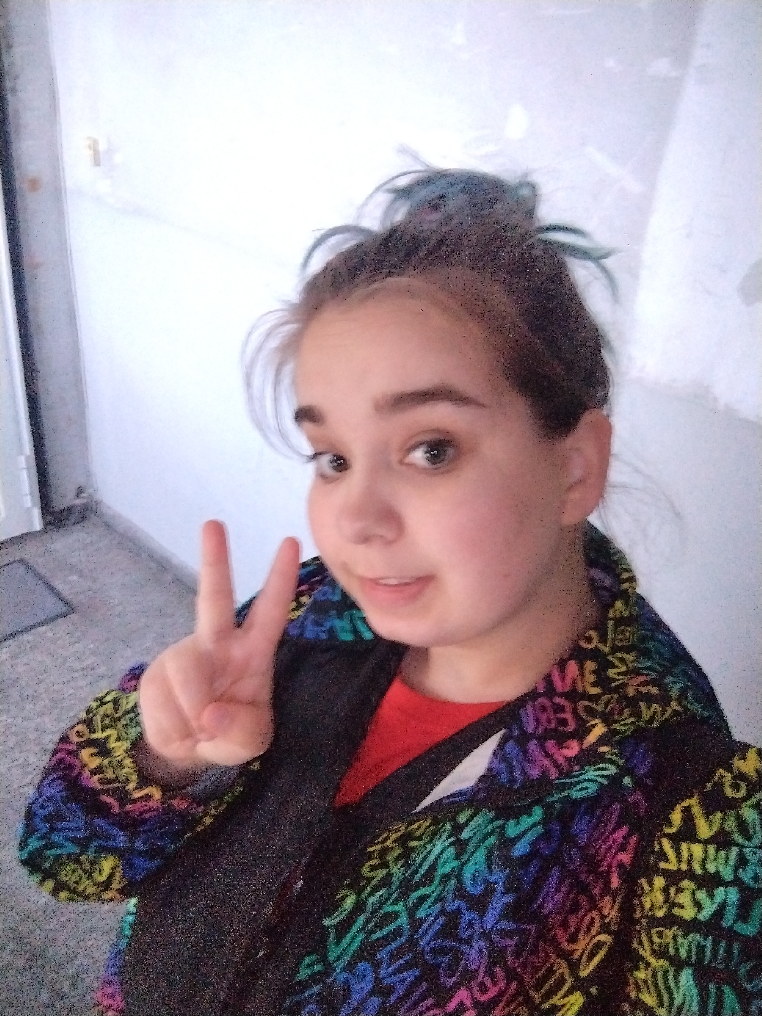 A young woman is taking a selfie in what appears to be an indoor hallway. She is holding up a peace sign with her left hand. The woman has light skin, brown hair with some blue streaks styled in a messy bun, and brown eyes. She is wearing a multi-colored jacket with a pattern of letters and words in rainbow hues over a red shirt.
The background consists of a white wall and a grey tiled floor. A closed white door is visible on the left side of the frame. The lighting is slightly dim.
There is no information in the image to suggest that this photo was taken in Plovdiv, Bulgaria.