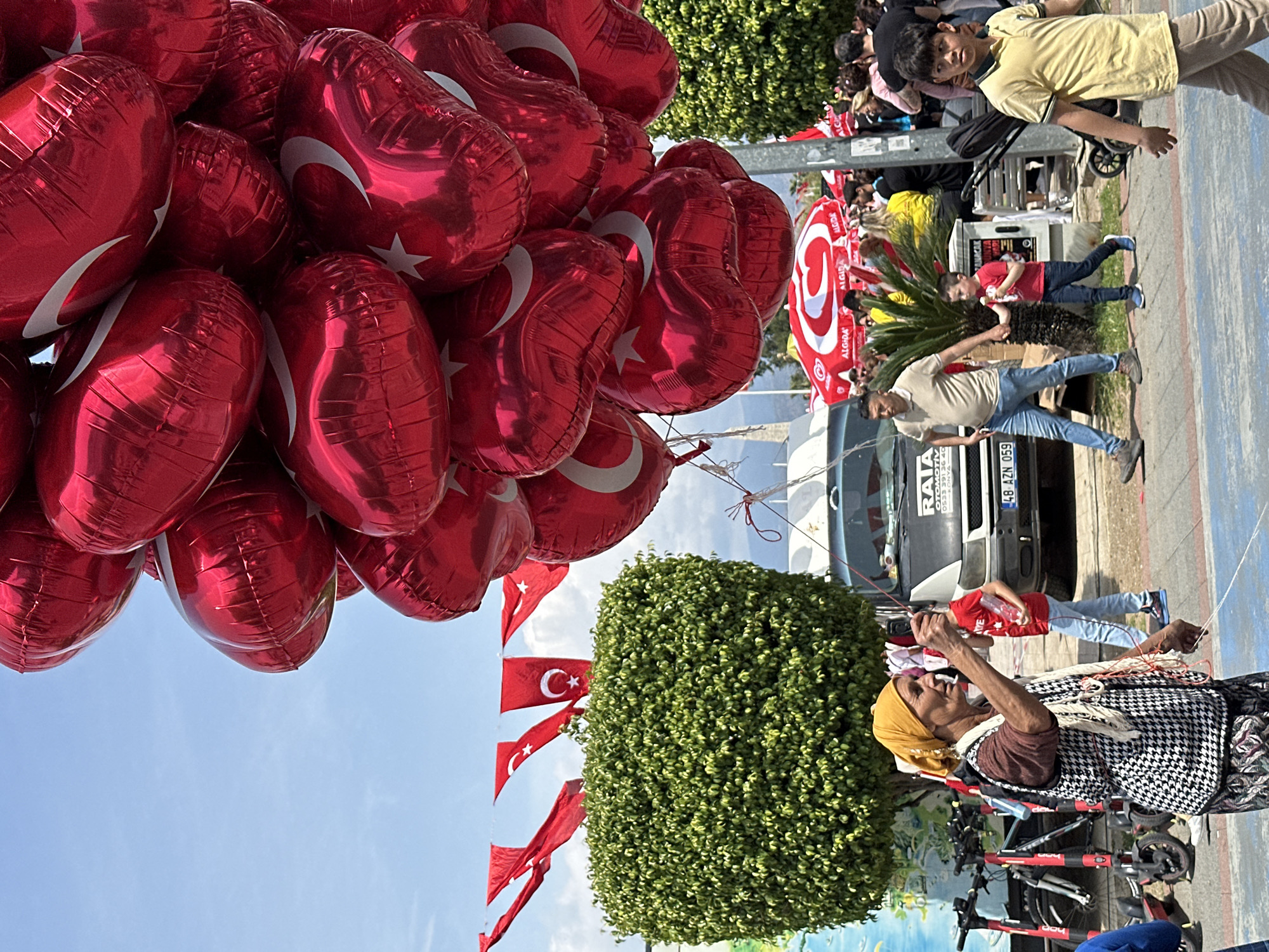 The image captures a vibrant outdoor scene in Fethiye, Turkey, under bright daylight. The foreground is dominated by a large cluster of glossy, red, heart-shaped foil balloons, each adorned with the white crescent moon and star motif of the Turkish flag. The sky is clear blue, visible between and above the balloons.
In the midground and background, various people are visible, suggesting a public gathering or celebration. A prominent figure is an elderly woman wearing a yellow headscarf and a black and white patterned vest, appearing to hold strings connected to balloons or other items. She is standing near several parked bicycles and scooters. Behind her, a white van or truck is partially visible, displaying the Turkish license plate "48 AZN 059," which corresponds to Muğla Province where Fethiye is located.
Other individuals include a young boy in a yellow shirt looking towards the camera, and another boy in a red shirt standing near a green bush. Several red Turkish flags are also seen hanging in the background, near more green foliage. Red and white striped umbrellas, some bearing the "ALGIDA" brand name, are visible further back, indicating a commercial or event setup. The overall atmosphere suggests a festive occasion or a nationalistic event.