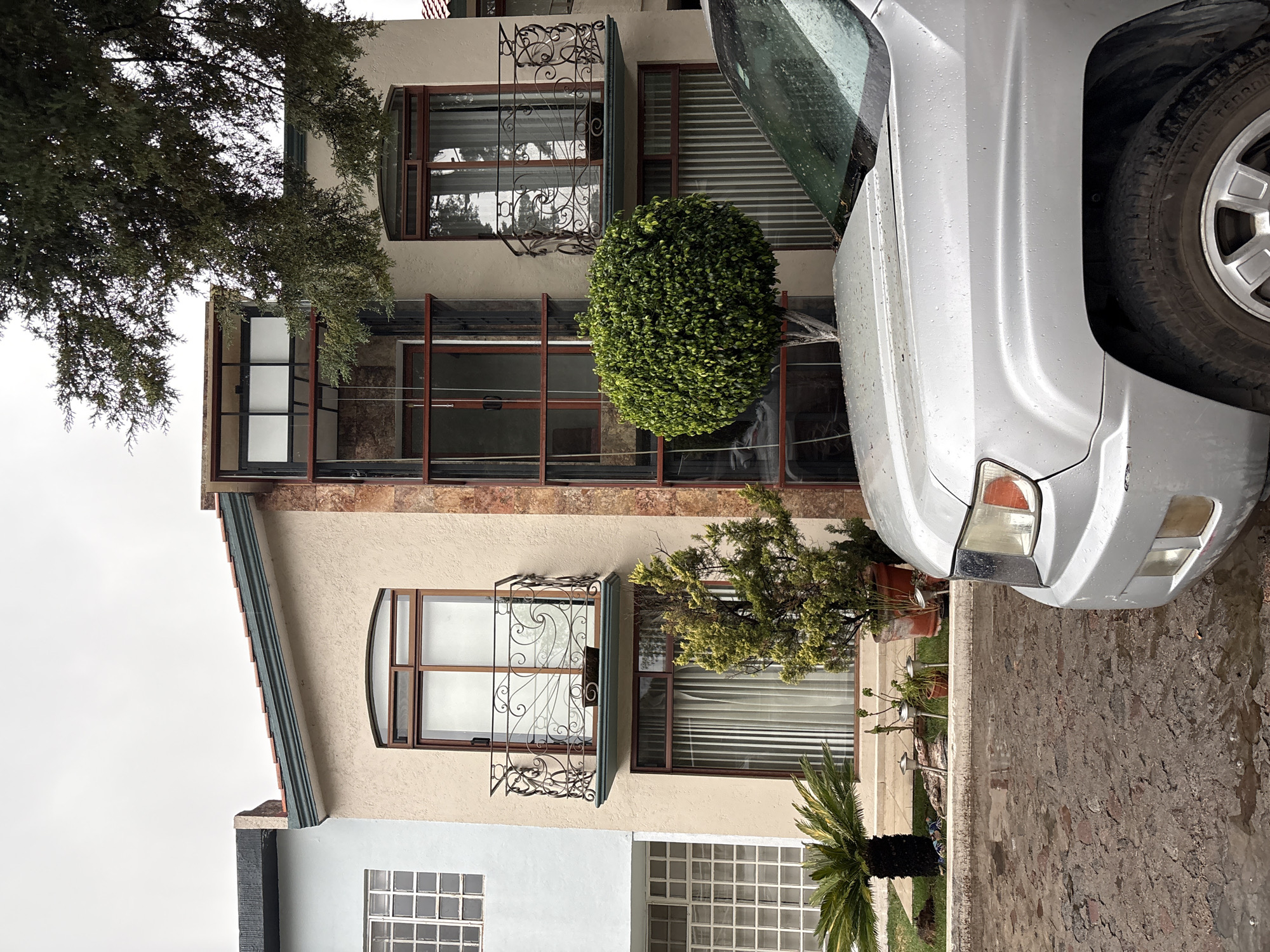 A multi-story building in San Francisco Totimehuacán, Mexico, exhibits a light beige facade with brown window frames. The windows on the upper floors feature ornate wrought iron decorative elements. A green, spherical bush is positioned on one of the floors. The front of a silver car is prominently visible on the right side of the image. A stone wall borders the property. A small palm tree is visible in the front yard. Overcast sky conditions.