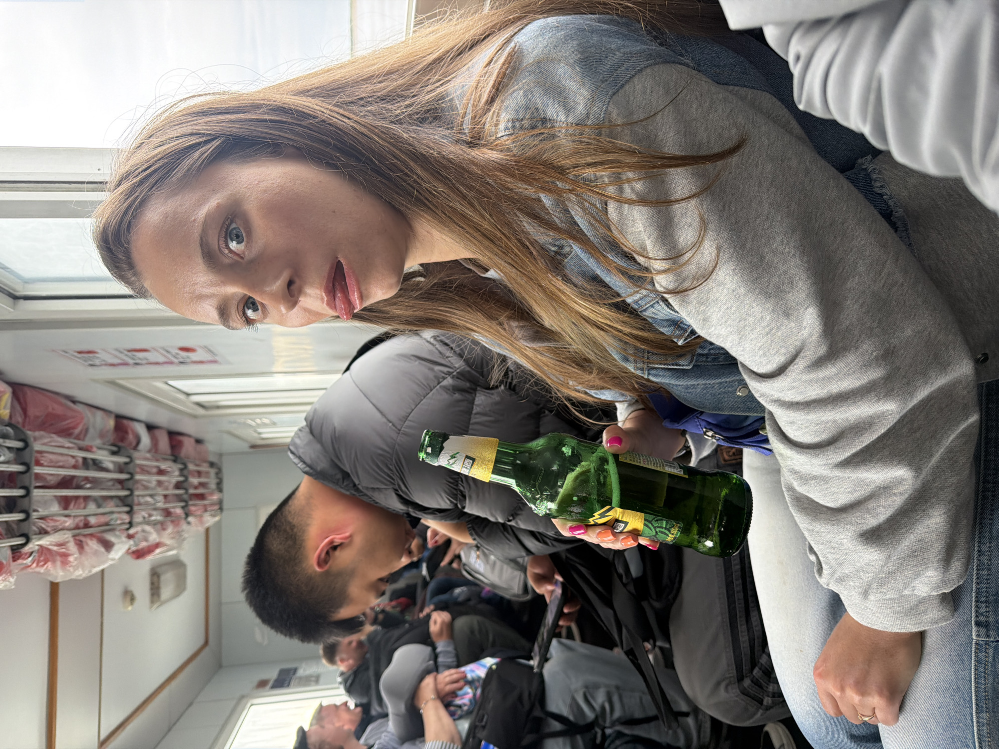 An eye-level shot captures a woman looking playfully at the camera, her tongue slightly out, inside what appears to be a passenger cabin of a ferry or similar public transport. She has long, light brown hair and is wearing a light gray sweatshirt over a denim jacket, paired with light-colored jeans. In her right hand, adorned with bright pink and orange nail polish, she holds a green bottle, likely beer, with a prominent yellow label displaying Asian characters. A gold ring is visible on her left ring finger.
Behind her, a man in a dark puffer jacket is partially visible, facing away from the camera. Several other passengers are also visible, seated or standing, filling the compartment. The setting features rows of shelves overhead, stacked with numerous red-packaged items, possibly emergency equipment. Daylight streams through windows along the side of the cabin. Some red symbols or text are visible on a white panel beneath a window. The overall scene conveys a casual travel environment during the day.