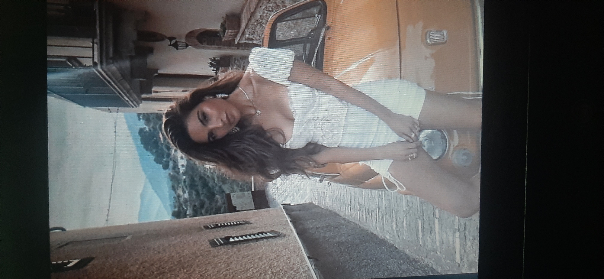 The image shows a woman in a white dress leaning against an orange car. She has long, dark, wavy hair and is wearing earrings and a necklace. The car appears to be a vintage Fiat. The background suggests a European town with stone buildings and a hilly landscape. There is no indication of Yessentukskaya, Russia, in the image.