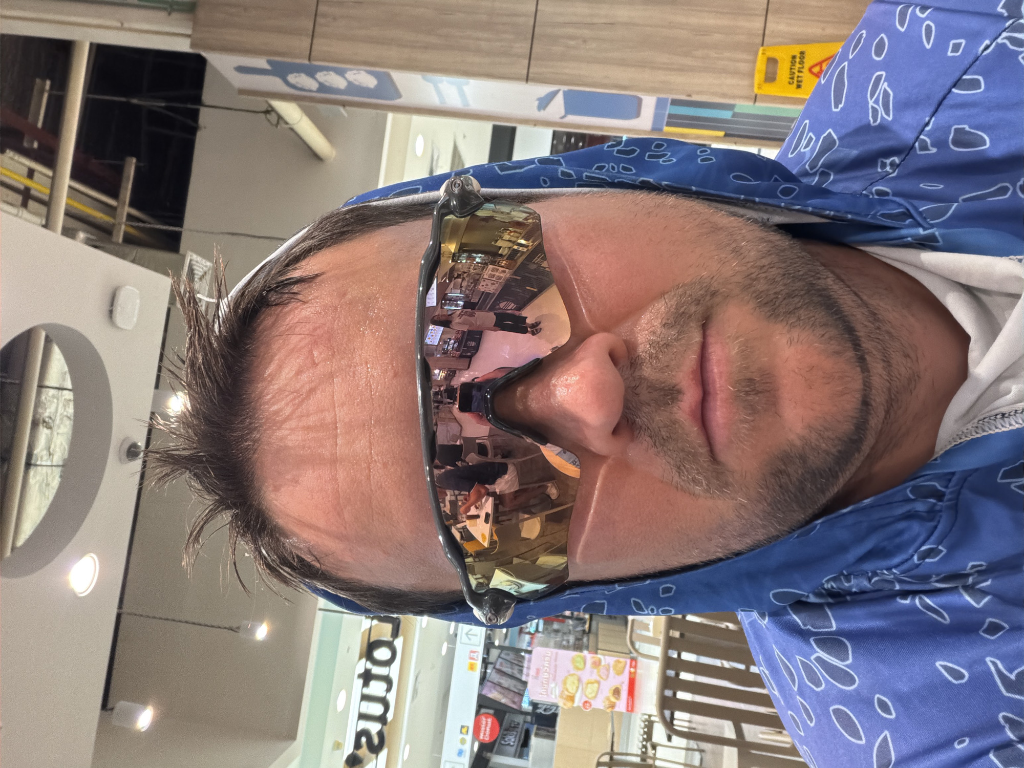 The image is a close-up selfie taken indoors, likely at a food court or a commercial establishment. The subject, a man with short, messy brown hair and stubble, is wearing sunglasses with reflective lenses and a blue hooded jacket with a white bandana-like pattern. The sunglasses reflect the environment, showing a food stall with people ordering and eating at tables. In the background of the main image, a sign with Thai writing is visible, along with seating areas. There are also overhead lights and ceiling structures suggesting an indoor public space. A yellow "CAUTION WET FLOOR" sign is visible on the right side of the image. The location is identified as a commercial area in Ban Bang Rak, Thailand, which implies a shopping mall or a similar venue.