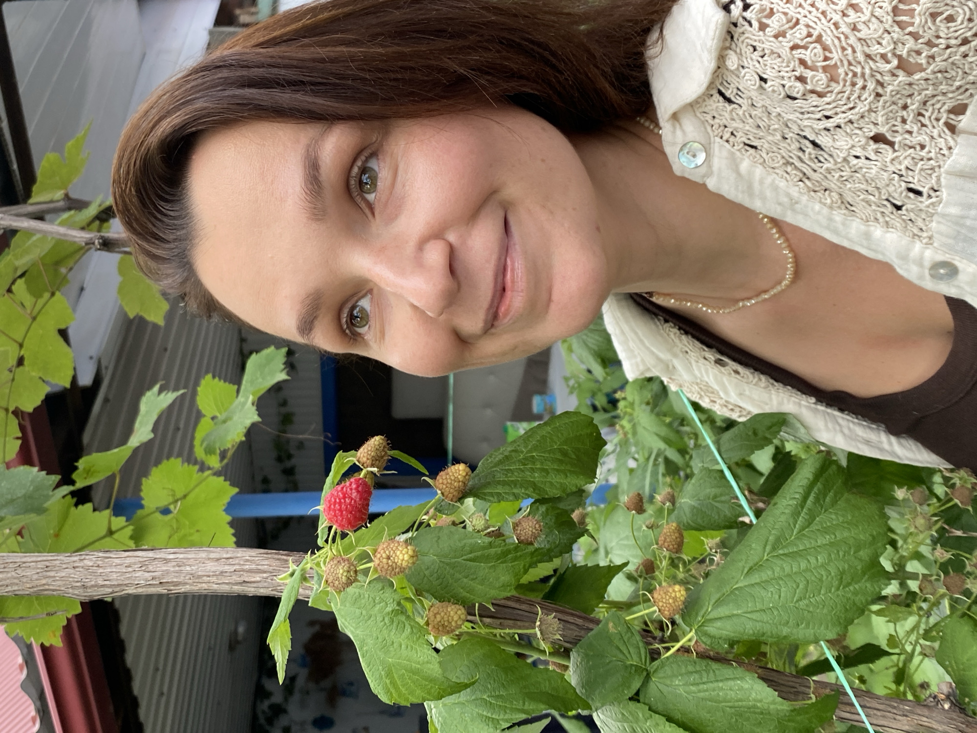 A woman is looking up and to the right with a slight smile, her face partially illuminated. She has brown hair and her eyes are light green. She is wearing a light-colored crocheted cardigan over a dark top and a pearl necklace. To her left is a raspberry bush with green leaves and several unripe raspberries, as well as one ripe red raspberry. The background is blurred but appears to be an outdoor setting with some structures and foliage, possibly in a backyard or garden. The lighting suggests it is daytime.