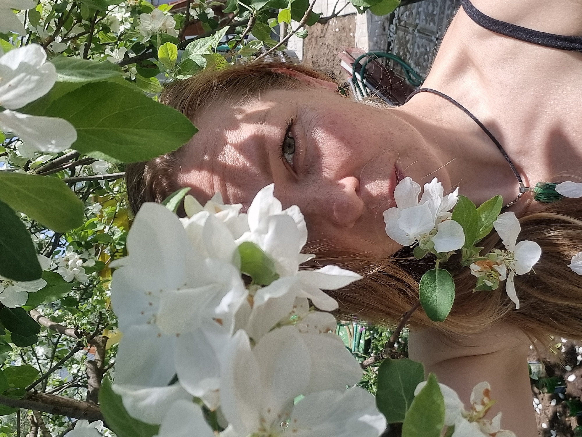 A photograph depicts a light-skinned person with freckles, viewed from a low angle amongst white flowers and green foliage, possibly in Chaikovsky, Russia. The person’s face is partially obscured by the flowers and shaded by sunlight filtering through the leaves. The person's eye is visible, appearing to be greenish. The person has light-colored hair. A black choker is visible around the person's neck. The background includes blurred foliage and branches.