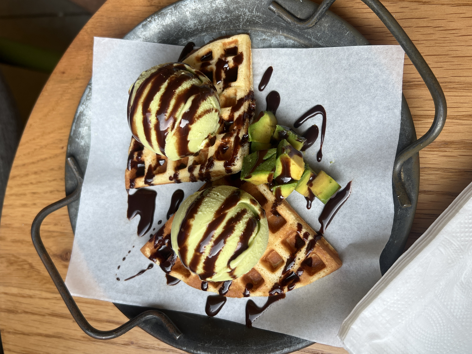 This overhead shot captures a serving of dessert or brunch on a rustic metal tray, placed on a light brown wooden table. The main subjects are two triangular portions of waffle, each topped with a generous scoop of pale green ice cream, possibly avocado or pistachio flavored. Both the ice cream and waffles are generously drizzled with dark chocolate syrup, creating striking visual lines and pools.
Between the two waffle pieces, a small pile of freshly cubed avocado is visible, also adorned with chocolate drizzle, adding an unusual savory-sweet element to the dish. The food is presented on a sheet of white parchment paper, which lines the metal tray. The tray itself is dark, likely cast iron or a similar metal, with visible texture and thick, curved handles on its sides. To the right of the tray, a neatly folded white paper napkin rests on the wooden table.
The lighting is bright and even, suggesting a daytime setting. There are no people or discernible text in the image. The overall scene conveys a casual and inviting atmosphere, likely from a cafe or restaurant in Nha Trang Ward, Vietnam, where this unique dessert combination might be offered.