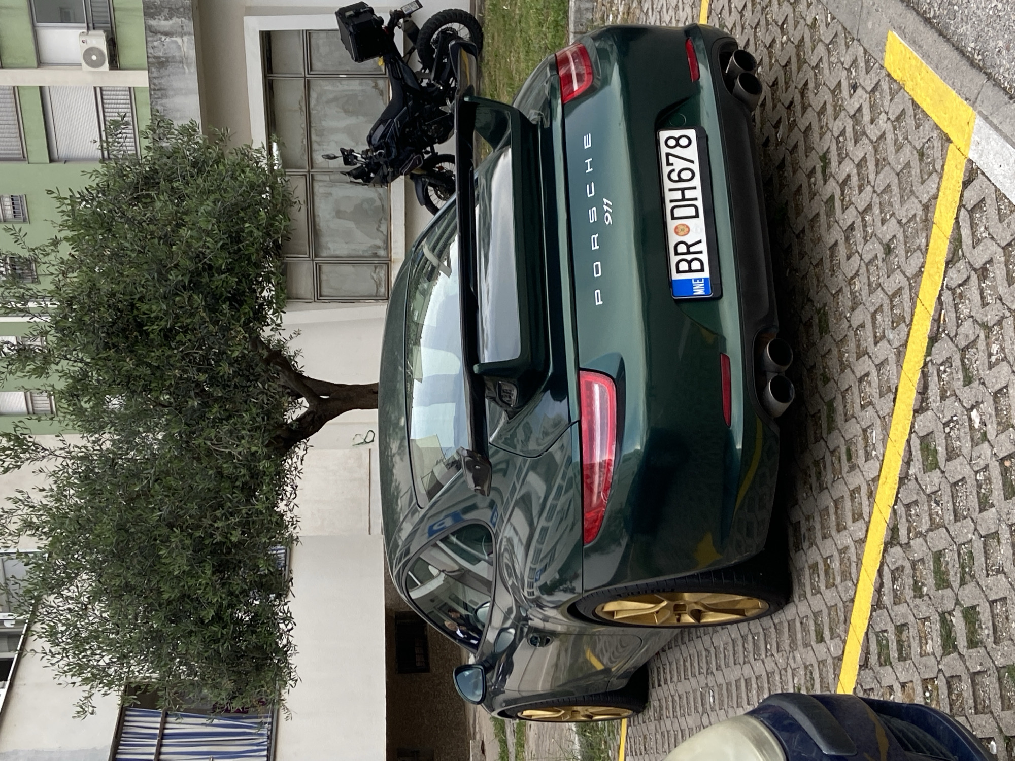 A green Porsche 911 with gold wheels is parked in a parking spot marked with yellow lines in Bar, Montenegro. The license plate reads "BR DH678" and indicates that the car is from Bar. A motorcycle is parked behind the car. A tree is next to the car. A building is behind the tree.