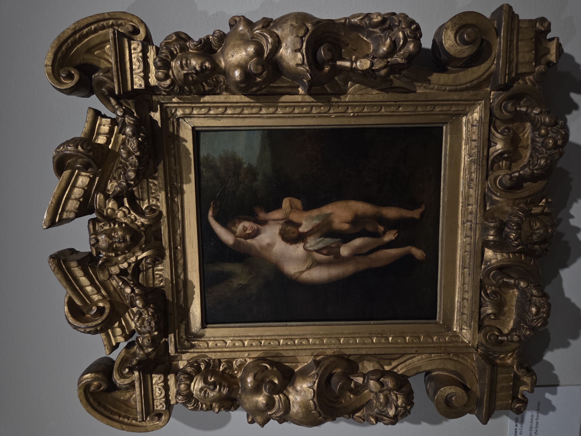 The image shows a painting in a highly ornate, gilded frame. The painting itself depicts two figures: a nude female figure with her arm raised, and a winged, nude cherubic figure embracing her. The background of the painting is dark and indistinct, with hints of trees and sky. The frame is elaborate, featuring sculpted figures of female busts and cherubic faces, along with scrolling acanthus leaves and other decorative motifs. The painting and frame are displayed on a light grey wall, with shadows cast to the lower left. A small white label with black text is partially visible in the lower left corner, indicating that the artwork is located in Malmö, Sweden.