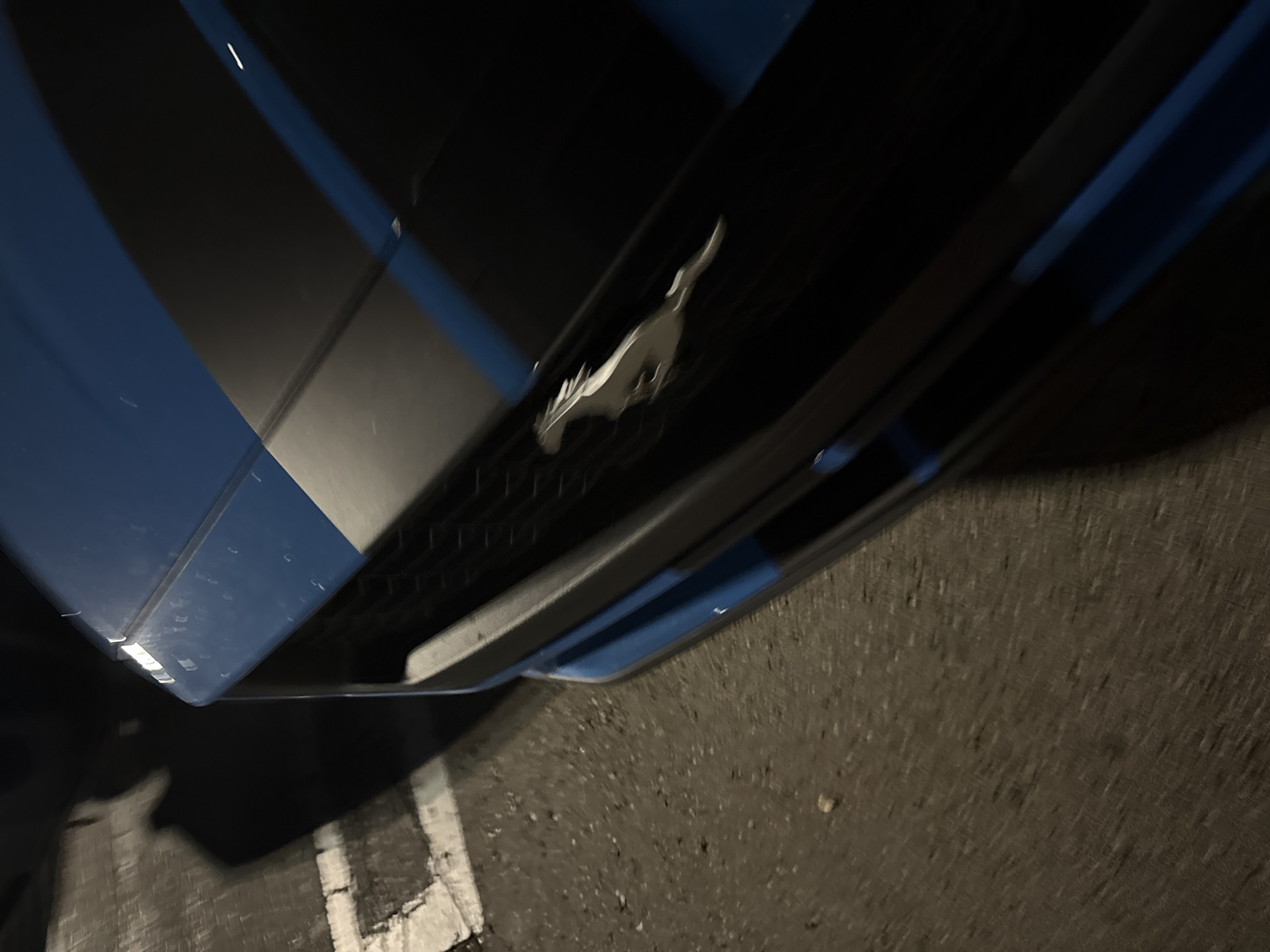 An eye-level shot captures the front of a blue Ford Mustang, illuminated under low-light conditions, suggesting nighttime or a dimly lit area. The iconic silver running horse emblem is prominently displayed on the car's dark, honeycomb-patterned front grille. Above the grille, a section of the car's blue hood is visible, featuring a wide, dark stripe, indicative of racing stripes. The car's bumper is also dark, reflecting some light.
The ground beneath the car appears to be asphalt or a similar paved surface, with faded white painted lines visible on the left and bottom right, suggesting the vehicle is in a parking lot or on a street. The overall lighting creates strong contrasts between the reflective surfaces of the car and the dark background. No people are visible, and no activity is occurring other than the stationary presence of the car. The image has a slightly tilted or rotated perspective. There is no visible text. The car appears to be clean.