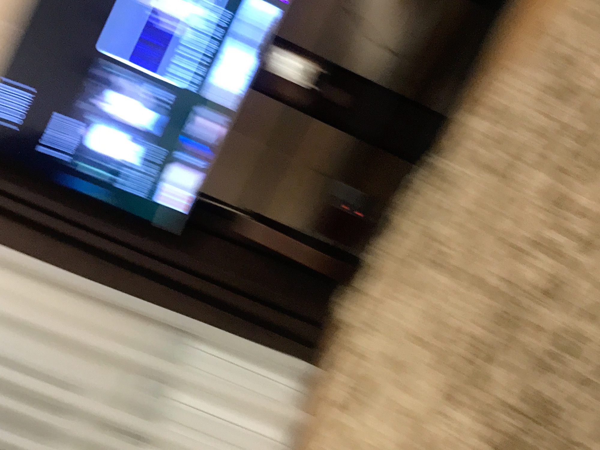 The image is a blurry, close-up shot of a television screen showing an interface with blue and white elements. The TV appears to be placed on a piece of furniture with a dark brown wooden frame and a white front panel. To the right of the TV, there is a blurry patch of tan carpet. The location is unknown and not discernible from this image.
