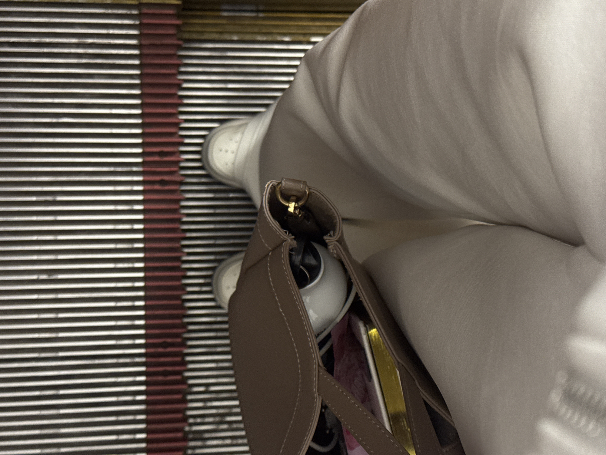 In this overhead shot of an escalator, a person's legs are visible from the knees down, clad in light-colored pants. They are wearing white sneakers. A brown handbag rests on the person's lap, with the opening facing upwards. Inside the bag, various items are visible, including what appears to be a white spherical object, possibly a camera or speaker, and some colorful papers or books. The escalator steps are a mix of silver and red-brown ribbed metal. The scene suggests a moment of stillness on a moving walkway, possibly within a shopping mall or transit station.