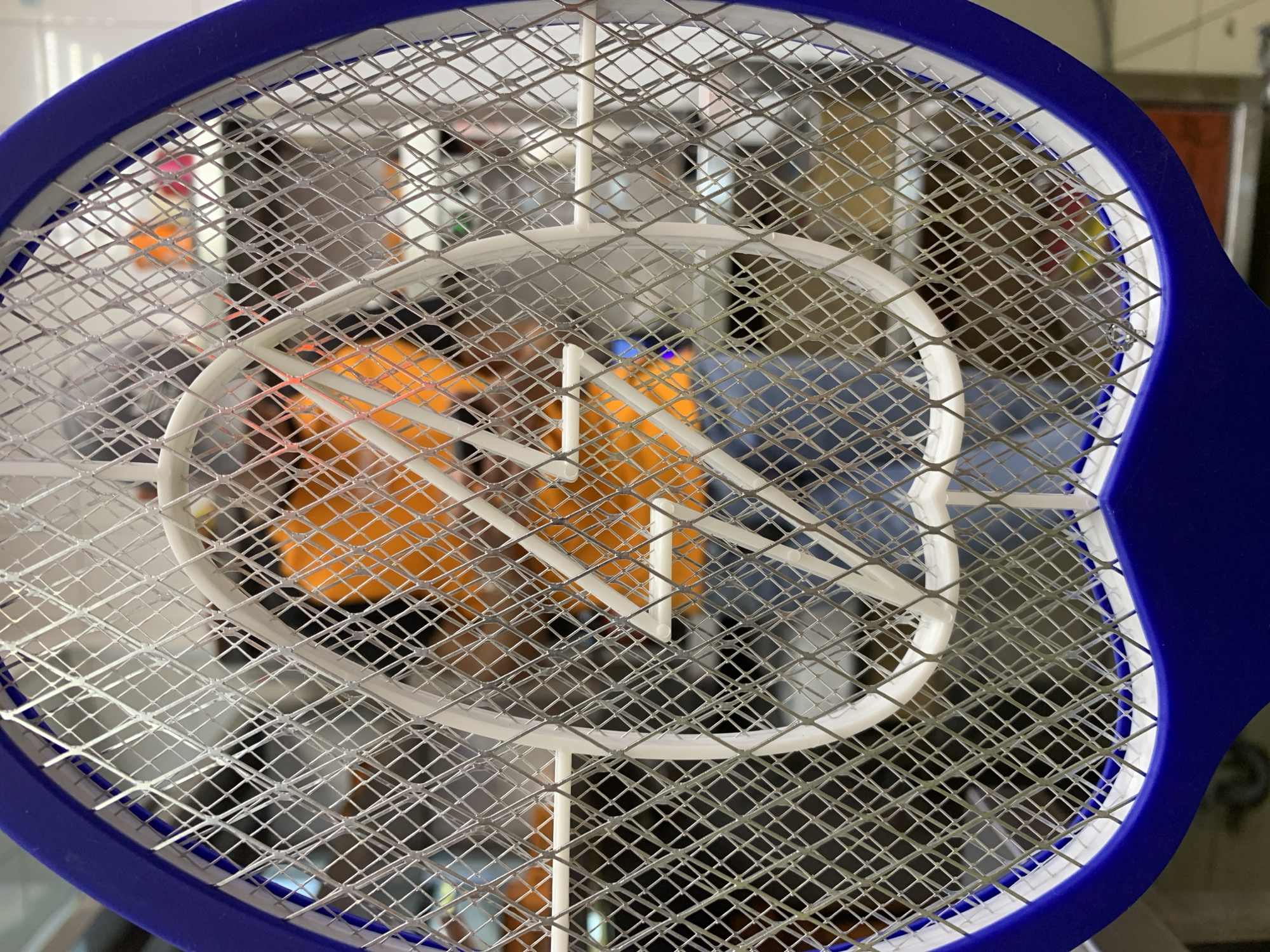 This media file is a close-up, low-angle shot of a blue and white electric mosquito swatter. The swatter has a mesh grid and a white oval frame. In the background, blurred figures of people can be seen. One person is wearing an orange shirt, and another appears to be wearing a blue shirt. The overall setting seems to be indoors, possibly a room or a hallway, with indistinct structures and objects visible in the background. The lighting is not particularly bright, suggesting it could be daytime but not a sunny day, or it could be indoors with artificial lighting. There are no discernible signs, text, or specific time-of-day cues. The image focuses on the texture and metallic sheen of the mosquito swatter's grid. The blurred background figures suggest the swatter is being held or is in use, but no specific action is clearly depicted.