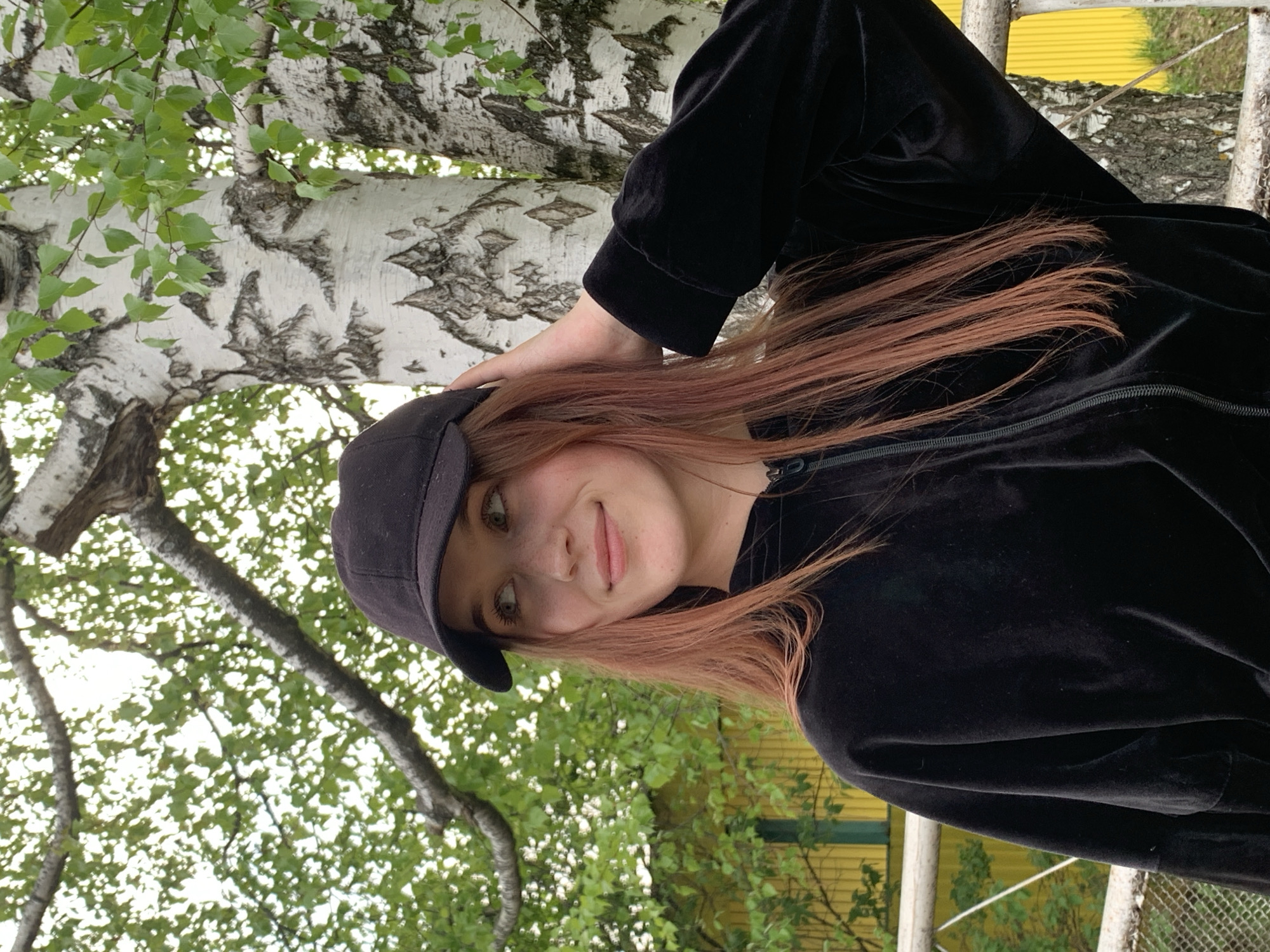 A young woman wearing a black cap and black velour zip-up jacket stands outside against a backdrop of birch trees with green leaves. She smiles slightly, and her hand rests on her head near her cap. In the background, a yellow building can be seen through the foliage. The location is Фоки, Russia.
