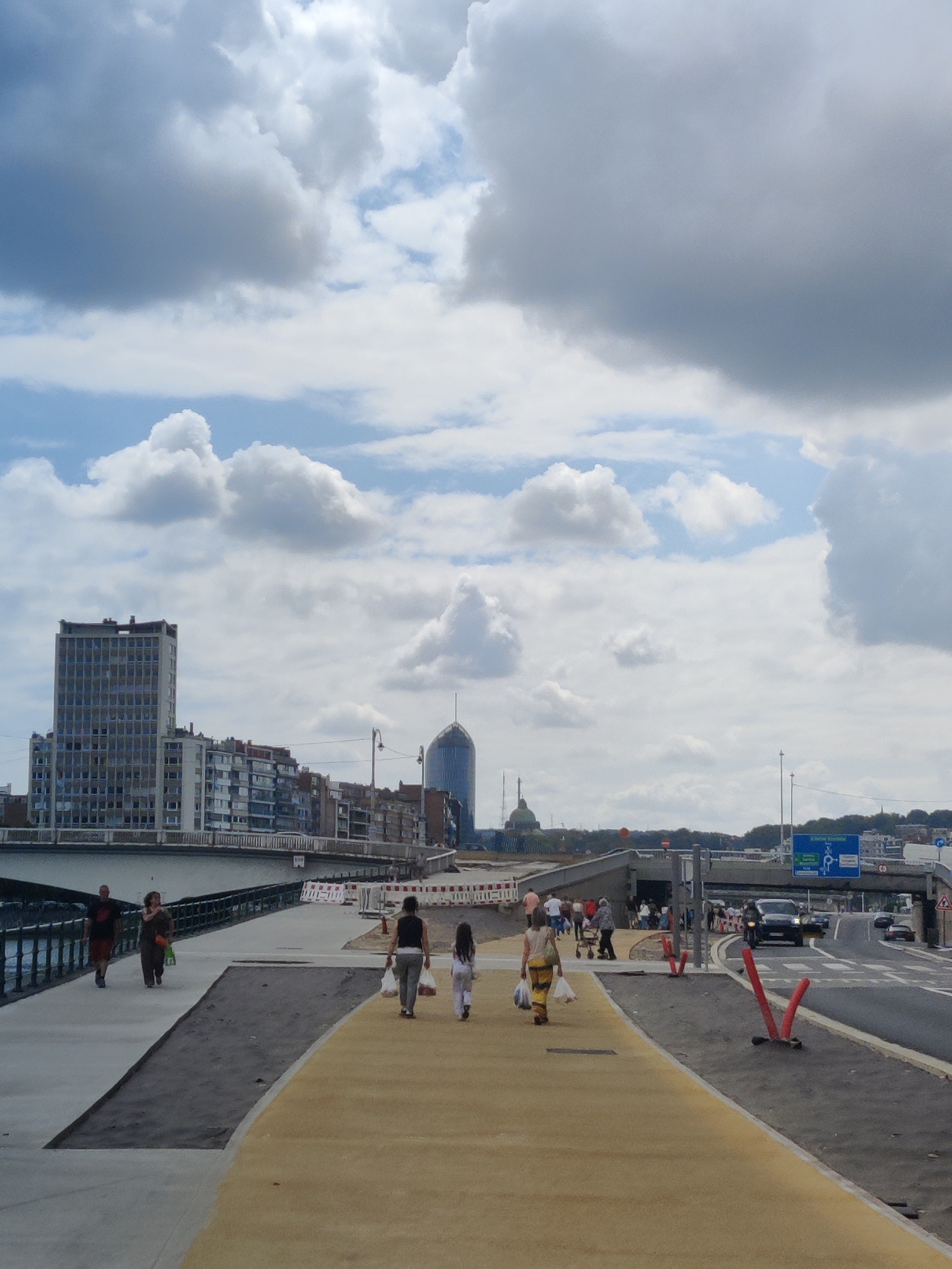 The image captures a scene on a pedestrian walkway alongside a road in the city of Liège, Belgium. In the background, a bridge spans the waterway, with several buildings lining the far bank, including a tall, rectangular building and a distinctive curved glass skyscraper. The sky is partly cloudy with dramatic formations.
On the walkway, several individuals are visible. On the left, a man and a woman are walking away from the camera. To the right, a woman in a striped dress carries shopping bags and walks towards the camera, followed by a young girl. Further ahead, another group of people are gathered near a construction zone marked by orange cones and barriers.
The walkway itself is under construction or recently completed, with sections of concrete and a yellow-tiled or painted path. The road adjacent to the walkway has traffic, including a dark-colored SUV and other vehicles, with traffic lights and road signs indicating directions. The overall environment suggests an urban setting with ongoing development.