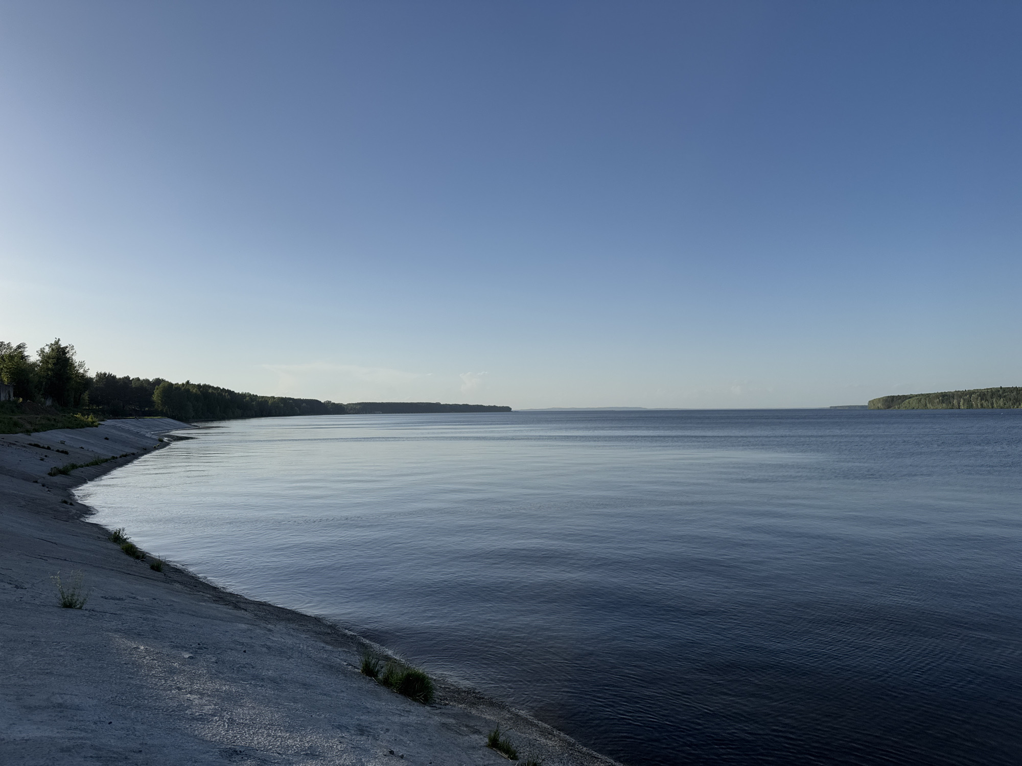 The image depicts a body of water, likely the Kama River, bordering the city of Chaikovsky, Russia. The foreground features a concrete embankment reinforced with grass, sloping down towards the water. A line of trees follows the shore along the left side of the image. Across the water, landmasses are visible in the distance, likely islands or the opposite shore. The sky is clear and blue. The water is calm with faint ripples. The overall scene is peaceful and quiet, likely captured during the morning or late afternoon, as the light is soft.