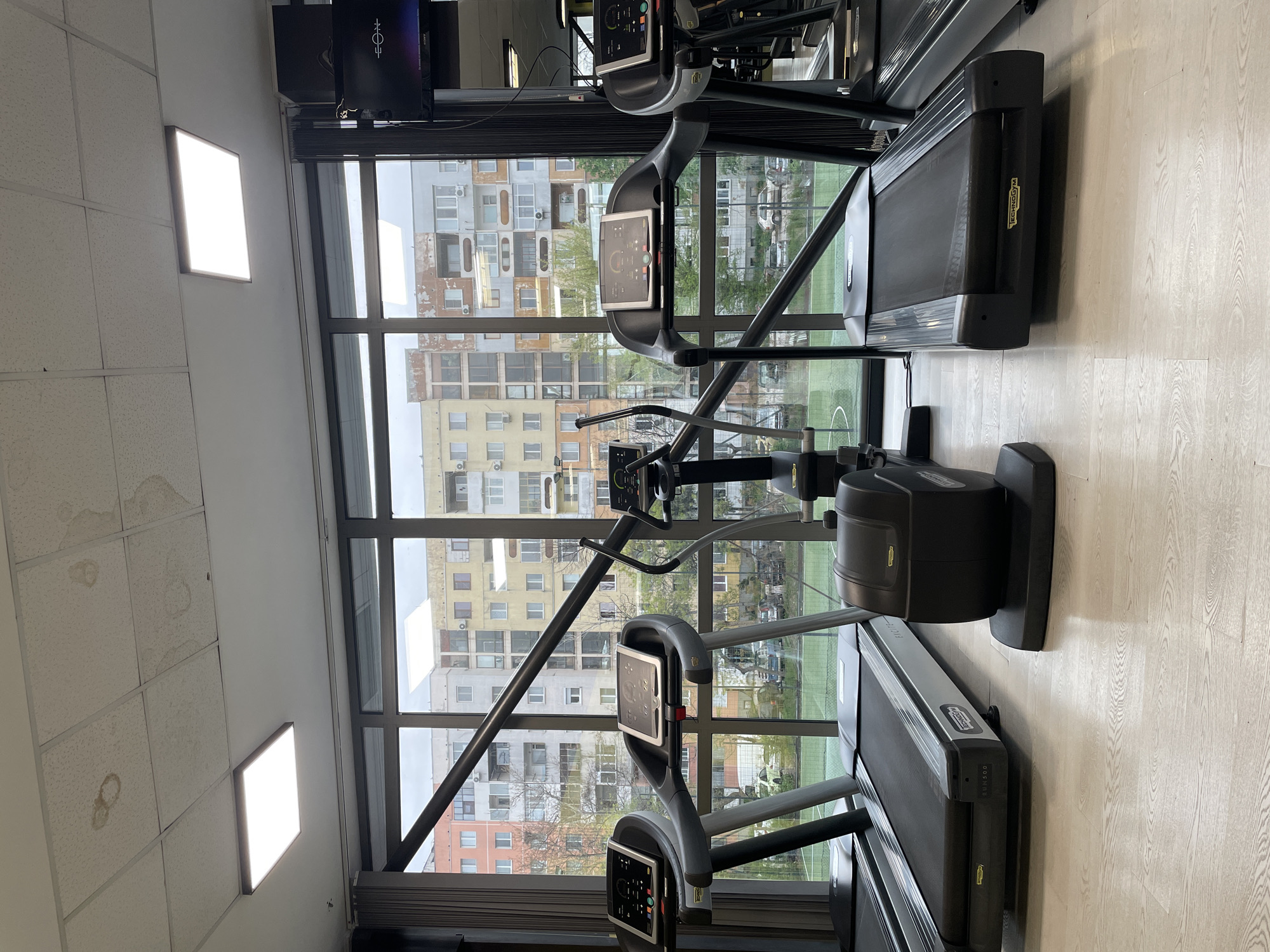 The image depicts the interior of a gym in Plovdiv, Bulgaria, with large windows overlooking an urban landscape of apartment buildings. Two treadmills face the windows, positioned on a light-colored, wood-look floor. A central elliptical machine sits between the treadmills. The gym's interior features a drop ceiling with recessed fluorescent lights. A television is mounted near the ceiling. The scene is focused on exercise equipment and the view outside, with no people visible. The outdoor view shows several apartment buildings with various colors and architectural styles.