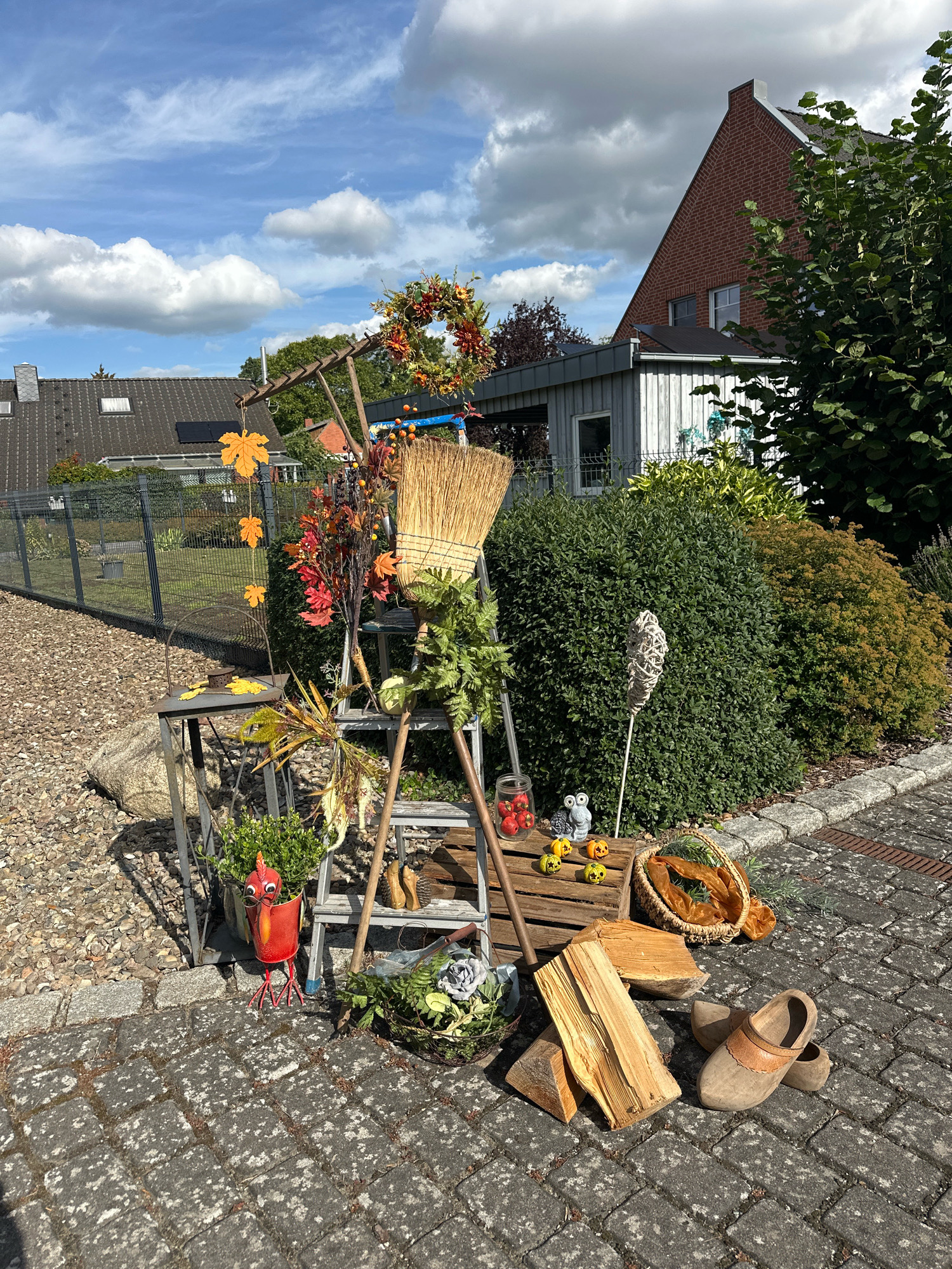 In the city of Samtgemeinde Thedinghausen, Germany, an outdoor display is set up on a paved area and surrounding gravel. The scene is decorated for autumn, featuring a variety of objects. A wooden ladder stands in the center, adorned with a broom, artificial autumn foliage, and a wreath of berries and leaves. In front of the ladder, a red metal rooster decoration is placed next to a potted plant.
To the left of the ladder, a small metal table holds decorative autumn leaves and a small metal object. Behind the table, a black metal fence encloses a gravel area.
On the right side of the ladder, a wooden crate holds a glass jar filled with red objects, small owl and mouse figurines, and mini pumpkins with carved faces. Next to the crate, a woven basket contains a shimmering orange fabric. Further to the right, a small stack of firewood is placed on the pavement, alongside a pair of wooden clogs.
The background shows a red brick house with a dark gray roof, a light gray shed with wooden siding, and lush green bushes and trees. The sky is blue with scattered white clouds. The overall impression is a festive autumnal display in a residential setting.