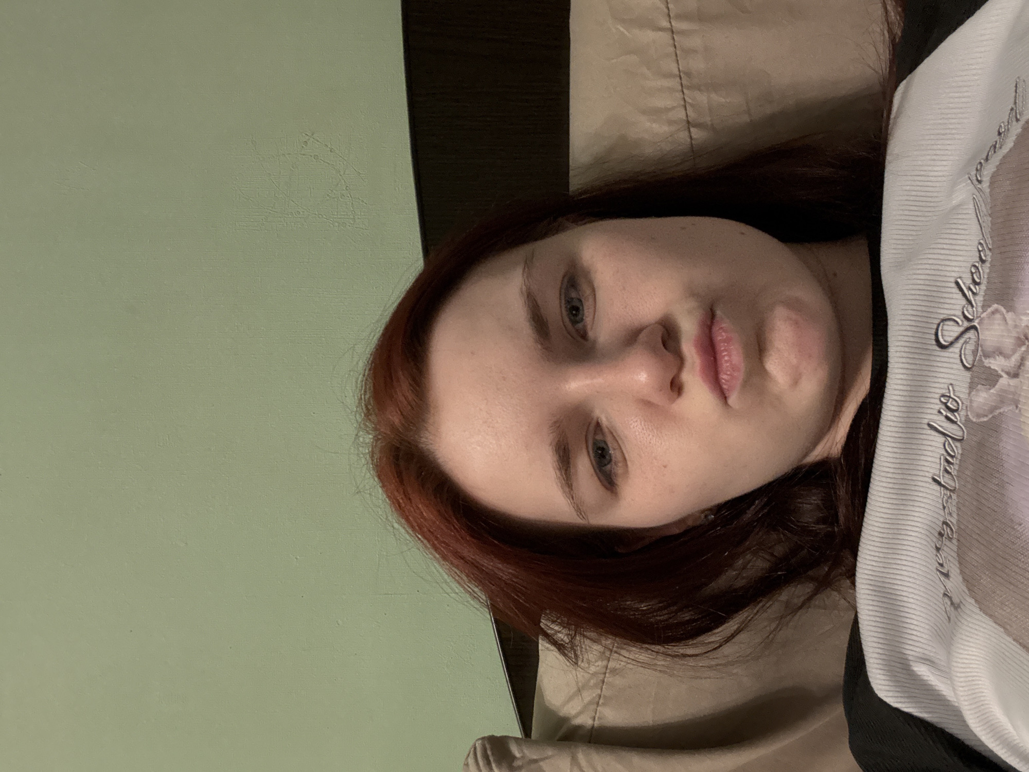 A young woman with reddish-brown hair is captured in a selfie, her face slightly angled and her lips pursed. She appears to be indoors, leaning against a light brown pillow and a dark headboard, with a pale green wall in the background. The woman is wearing a white t-shirt with black sleeves, featuring embroidered text. The image does not provide any information about the city of Sinyavino, Russia, nor does it depict any forensic or criminal activity.