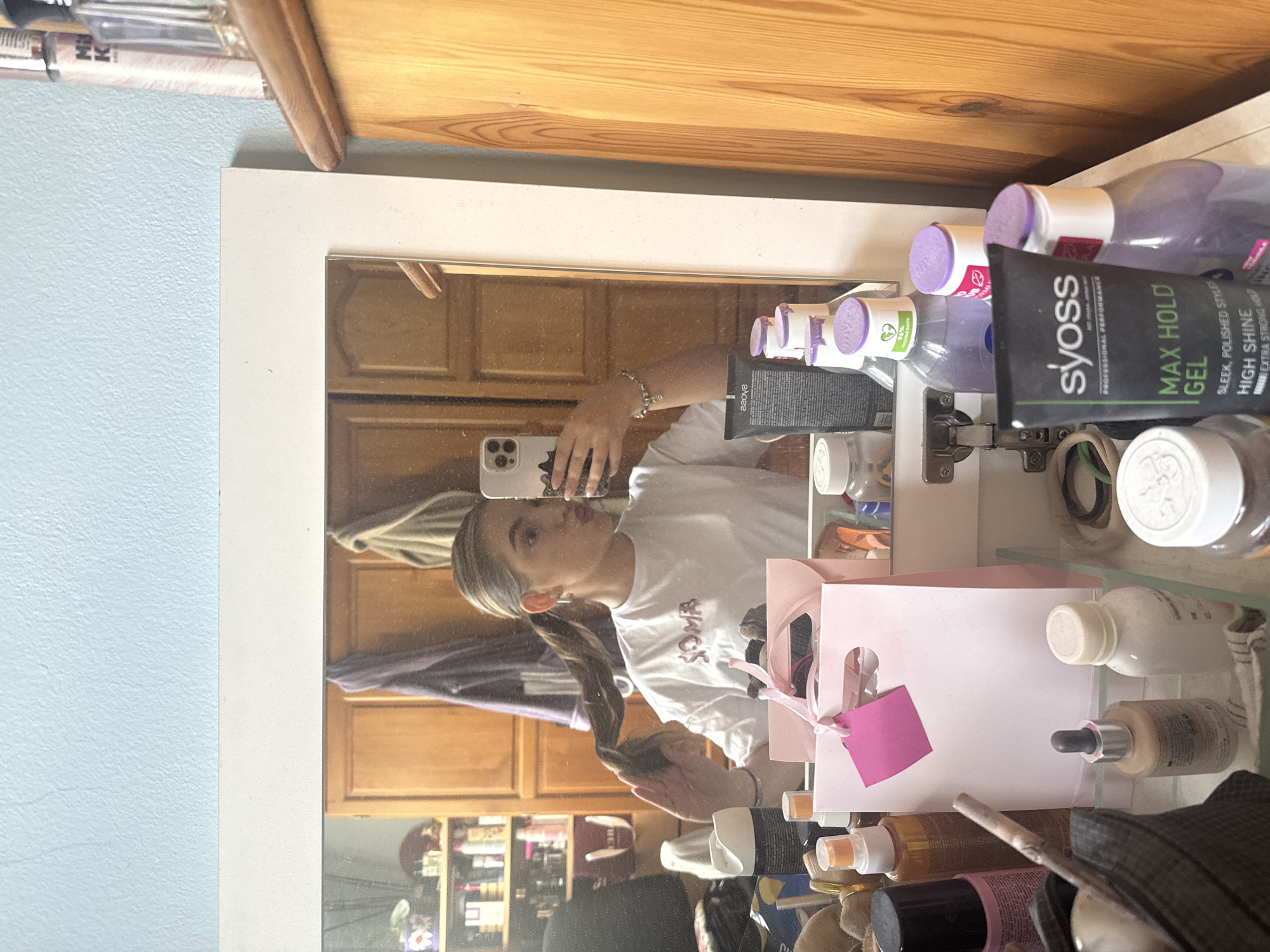 This media file shows a reflection of a young woman in a mirror. She has blonde hair tied in a ponytail and is wearing a white t-shirt with red lettering that reads "SOMA". She is holding a phone in her left hand, which is angled to take the picture. Her right hand is near her hair, and she appears to be adjusting it. A pink gift bag with a pink tag is visible next to her.
On the shelf in front of the mirror, various beauty products are displayed, including bottles of what appear to be hairspray or styling products, a tube of "syoss MAX HOLD GEL", and other skincare or makeup items. The setting appears to be a room with wooden cabinetry visible in the reflection behind the woman. The lighting suggests it might be daytime indoors, as there is ample light reflecting off surfaces. The overall mood seems casual and everyday.
Visible text includes "SOMA", "syoss", "PROFESSIONAL PERFORMANCE", "MAX HOLD", "GEL", "SLEEK. POLISHED STYLES", "HIGH SHINE", and "EXTRA STRONG HOLD". There is also text on bottles that reads "NIVEA" and "96% recycled plastic". The location context provided is Sofia, Bulgaria, which is not directly discernible from the image itself but is given as contextual information.