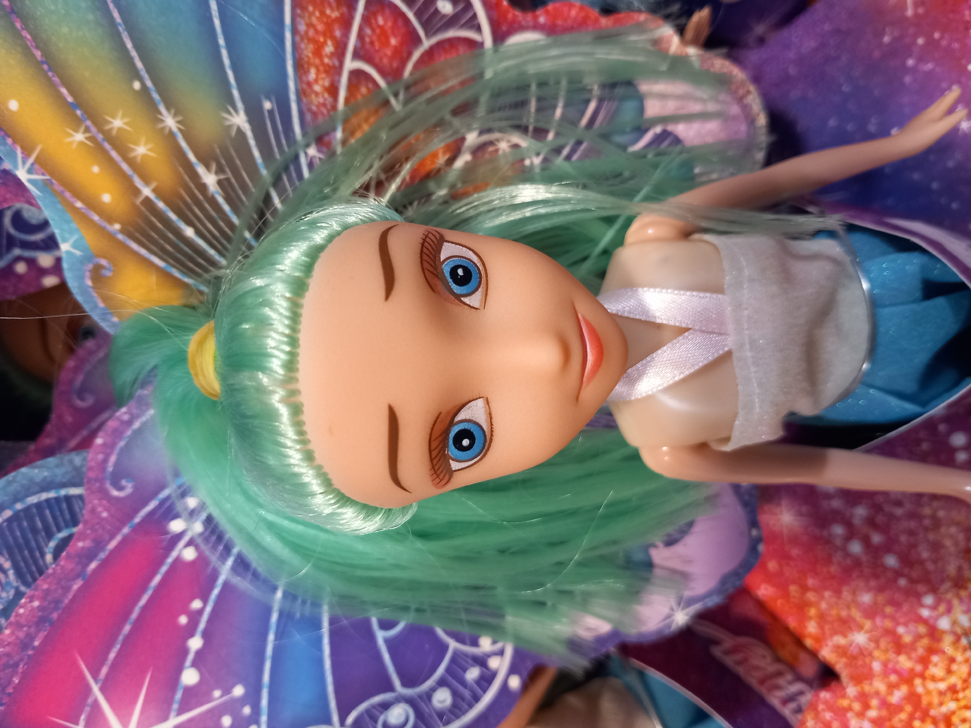 This media file shows a close-up, overhead view of a doll. The doll has long, light green hair and blue eyes. It is wearing a white top and a blue skirt. The doll is lying on a colorful background that appears to be made of patterned paper, possibly resembling fairy wings. The background features shades of purple, pink, yellow, and blue with sparkling white accents. One of the doll's arms is visible, bent at the elbow, with its hand slightly outstretched. The doll's face is tilted slightly upwards, and its mouth is slightly open. The overall impression is that of a toy fairy doll. There is no visible text in the image. The lighting suggests it might be an indoor setting, possibly a toy store or a child's room, but the exact location is unknown. No specific time of day or weather conditions are indicated.