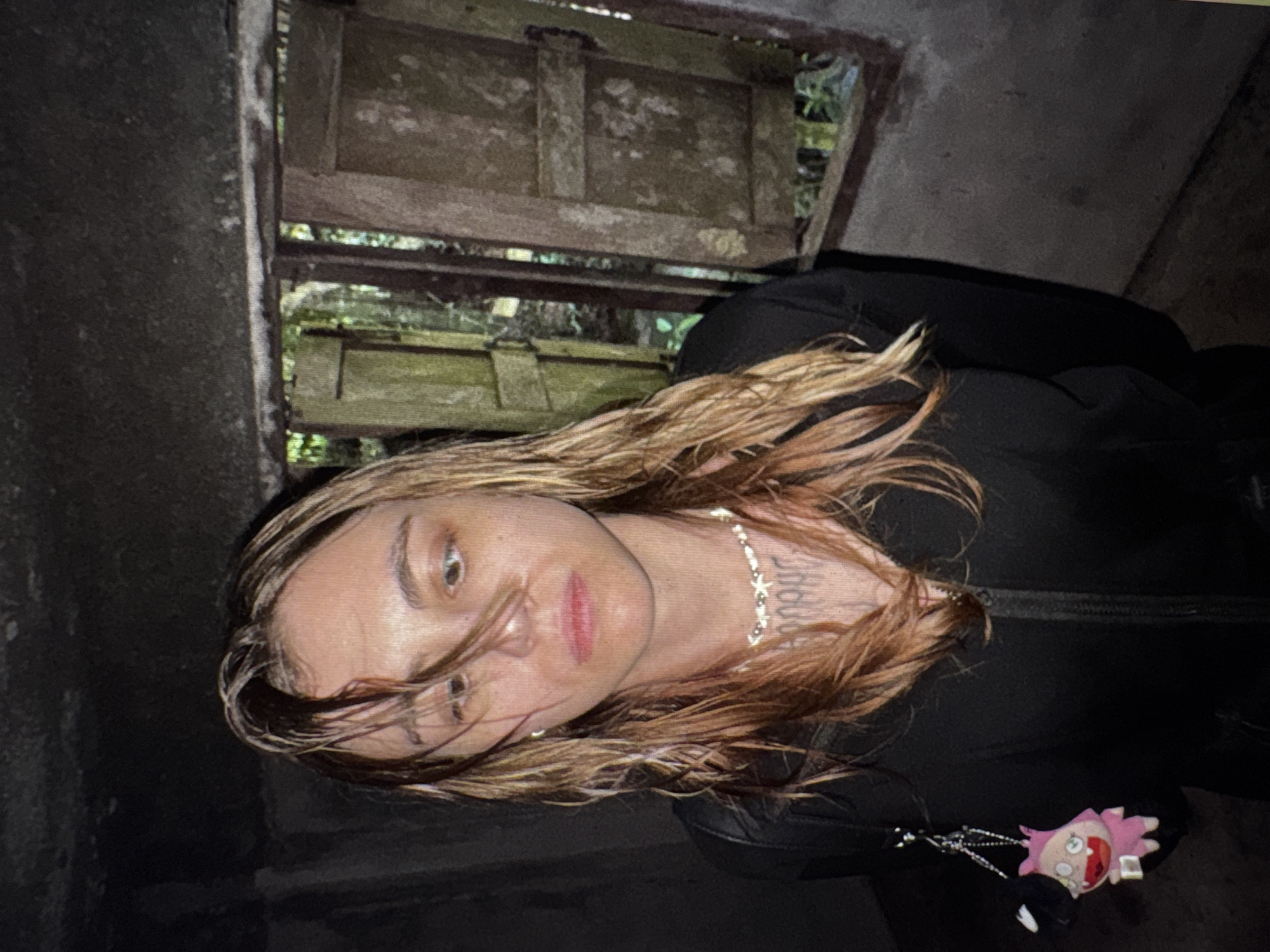 A young woman with wet, wavy blonde and brown hair stands in a dimly lit, rustic structure. She is wearing a black zip-up jacket over a necklace and a shirt or dress with visible tattoos on her chest. A pink plush toy with large eyes and a wide mouth hangs from a chain attached to her jacket. She has a pale complexion, with a slight blush on her cheeks and appears to be looking slightly downwards and to her left. Behind her, to the right, is a weathered wooden window with two shutters, through which a lush green forest can be seen. The walls of the structure are made of rough, dark concrete. The overall impression is one of an abandoned or dilapidated building in a natural, overgrown setting. There is no discernible information to place this scene within Moscow, Russia.