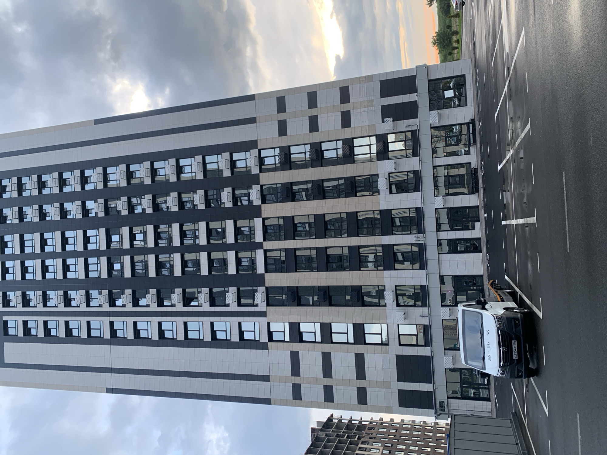 The image shows a modern, multi-story building with a facade of alternating dark and light panels and numerous windows. The building appears to be a residential or office complex, with a clean, contemporary design. In the foreground, a parking lot with white lane markings is visible. The lot is wet, suggesting recent rain, and the sky is overcast with patches of sunlight breaking through, indicating either early morning or late afternoon. A white van is parked in one of the spots. The license plate on the van reads "C315KP 126". The overall scene conveys a sense of urban development and a slightly damp, transitional time of day. The provided location context is Stavropol, Russia.