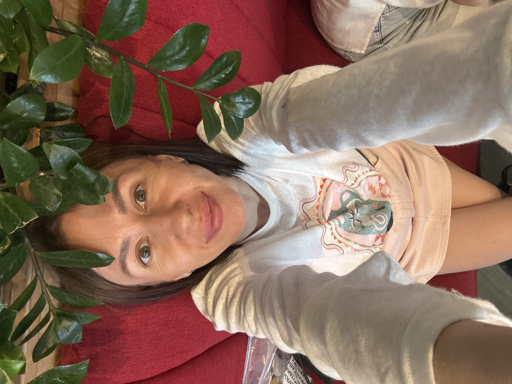 A woman with brown hair and brown eyes, wearing a white long-sleeved shirt with a graphic print and a light pink skirt, is lying on a red couch. She is holding a smartphone, taking a selfie. Green leaves from a plant are hanging over her head, obscuring the top of her forehead and the area above her. She is looking at the camera with a slight smile. The background is a textured red surface, likely the couch. The image is taken from a high angle, looking down at the woman. The environment appears to be indoors, possibly a home or a cafe. There is no identifiable information in the image that would place it in Izhevsk, Russia.