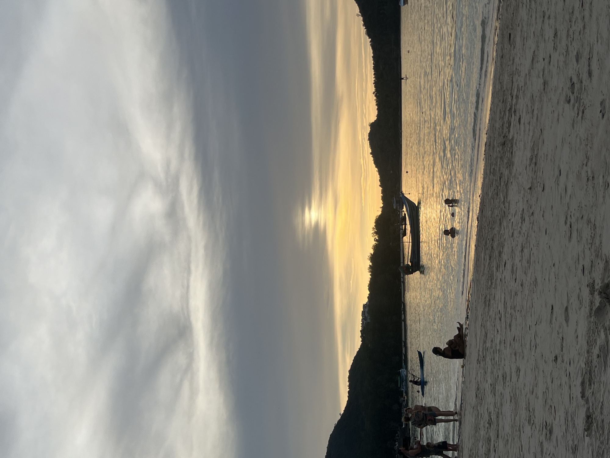 This is a full-frame, vertical, eye-level, medium shot photograph taken on a beach in the Ko Pha-ngan District of Thailand. The image depicts a twilight scene with a sandy beach in the foreground, the calm ocean in the middle ground, and a forested, hilly coastline under a cloudy sky in the background.
On the beach, several people are visible. To the left, a group of four individuals stand near the water's edge. Further to the right, a person is seated on the sand near the water. In the ocean, a few people are swimming or wading.
The water is a warm, golden hue, reflecting the setting sun which is partially obscured by clouds and positioned near the horizon. A long, narrow boat with an outboard motor is anchored in the water to the left of the frame's center. Another boat is visible further in the distance, as are several smaller vessels. The sky is dominated by gray and white clouds with streaks of yellow light from the setting sun. The overall mood is tranquil and serene.