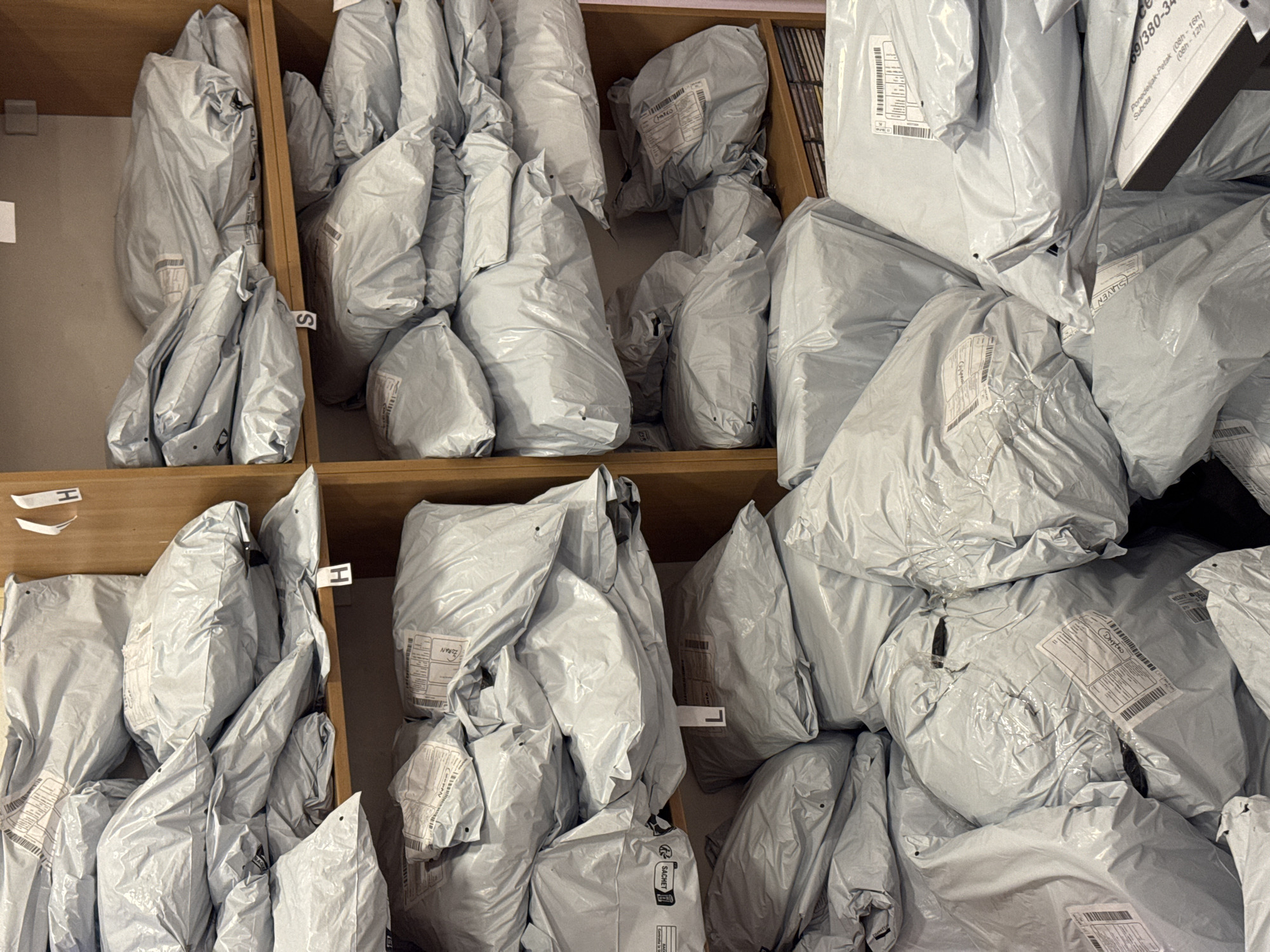 The image captures a static, indoor scene depicting numerous parcels and packages, primarily light gray plastic poly mailers, stored within wooden shelving units or cubbyholes. This arrangement strongly suggests a mail or parcel collection/distribution point, likely a post office or a delivery service center, given the context of Budva, Montenegro.
The packages vary in size and are densely packed into the compartments, with some overflowing. Many parcels bear white shipping labels with barcodes, tracking numbers, and recipient details, though most specific text on these labels is indistinct. Some cubbyholes are marked with simple, handwritten letters like "H" and "S," implying a sorting system.
Notable visible text includes names such as "MARCO", "ZORAN", "SLAVEN", and "DHYTRO" on various package labels, possibly indicating recipients or senders. The word "SACHET" is also visible. A particularly clear label in the upper right section displays operating hours: "Ponedeljak-Petak (08h-16h)" (Monday-Friday, 8 AM - 4 PM) and "Subota (08h-12h)" (Saturday, 8 AM - 12 PM), along with a numerical code "69/380-34." These hours are written in a South Slavic language, consistent with the Budva, Montenegro location context. No people are visible, and no active events are occurring; the image depicts a state of organized storage. The lighting suggests an indoor setting.