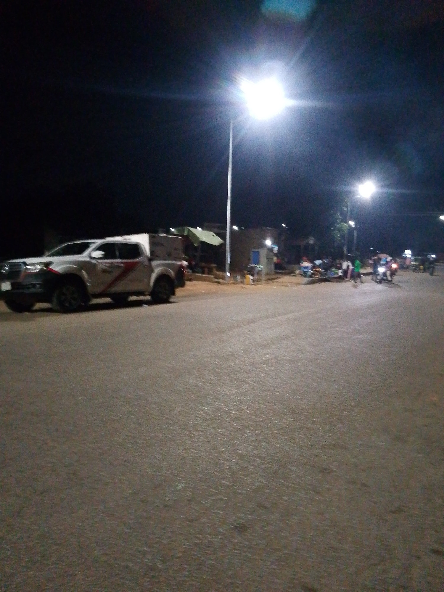 The image depicts a nighttime scene on a paved road, likely within a town or city in an unspecified location. A white pickup truck with a camper shell is parked on the left side of the road, facing away from the viewer. The truck has a red and grey decal on its side. Across the road, several people are gathered around a stall with a green umbrella. There are also motorcycles and what appears to be a small building or structure in the background. Streetlights illuminate the scene, casting a bright glow and creating lens flares. The overall atmosphere is dimly lit, suggesting it is late at night. The road surface is asphalt, and there are some shadows cast by the lighting.