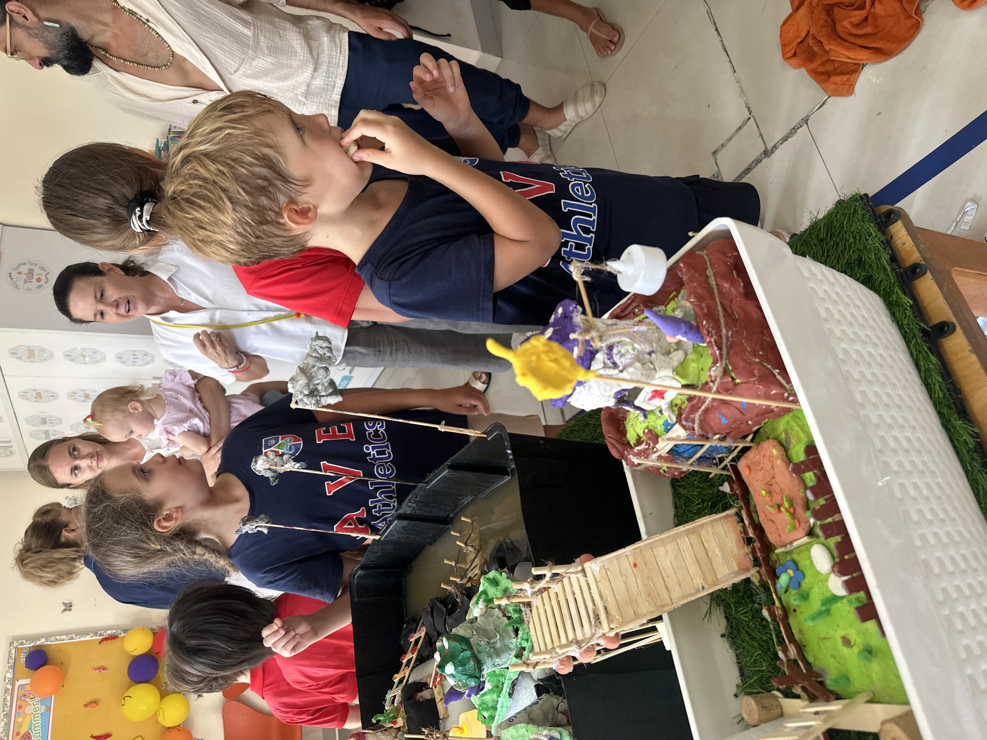 In a bright, indoor setting, possibly a classroom or community center in Nam Nha Trang Ward, Vietnam, a group of children and adults are gathered around a diorama. The diorama, constructed in a white, woven container, features a miniature landscape with a body of water, a wooden bridge, and colorful, handmade figures. Green artificial grass lines the base of the diorama, and small wooden fences create pathways.
A young boy in a navy blue t-shirt is in the foreground, looking towards the diorama and eating something small. Behind him, a young girl with braided hair, also in a navy blue t-shirt, is looking to her right. Another child in a red shirt stands to the left, partially visible.
Adults are interspersed among the children. A woman in a white shirt with a lanyard is speaking, looking towards the group. Another woman, holding a baby, stands behind her. A man with a beard and glasses, wearing a cream-colored open shirt, is on the right side of the image, looking towards the diorama.
The background shows a bulletin board with colorful decorations, possibly student artwork, and part of a whiteboard or cabinet with text and drawings. The floor is tiled with a beige and white pattern. An orange towel is visible on the floor to the right. The scene appears to be a school event or exhibition.