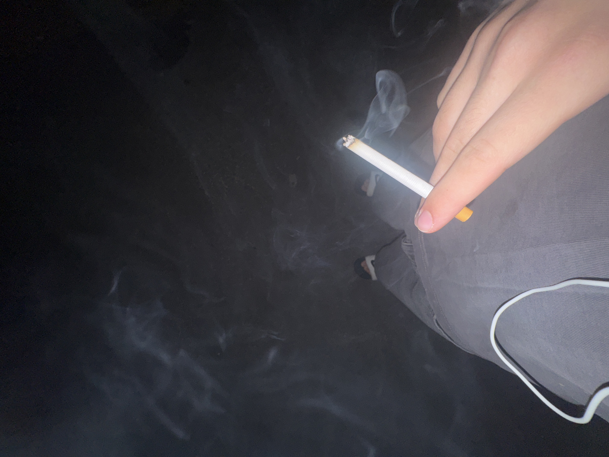 The image shows a close-up view of a person holding a lit cigarette, with smoke visible around it. The person is wearing dark gray pants and a white earphone cord is draped over their leg. The background is dark and out of focus, suggesting an outdoor or dimly lit environment. There is no specific visual information in the image to determine its location within the city of Trenčín, Slovakia. The scene is static, focusing on the act of smoking.